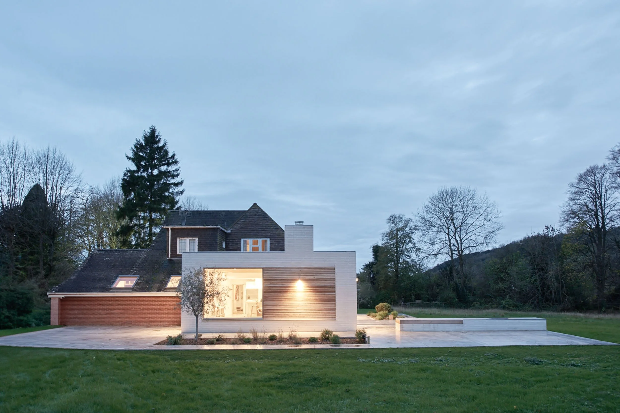 Modern house with large windows, lit from inside, situated in a landscaped yard with trees and mountains in the background during dusk.