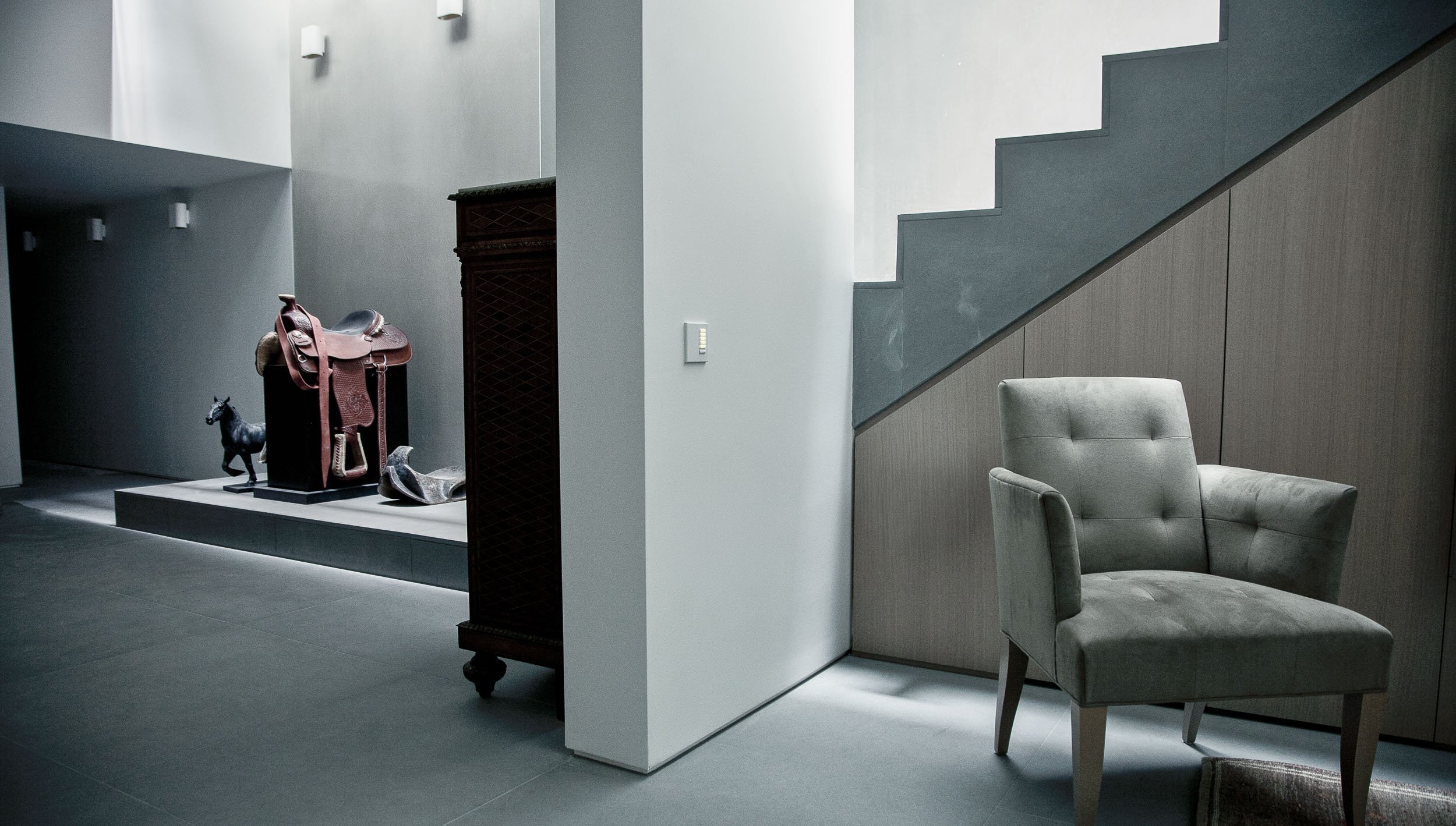 Modern, minimalist interior with a beige armchair near a dark wall and staircase, a display with a saddle and horse sculpture, and a wooden cabinet under soft lighting.