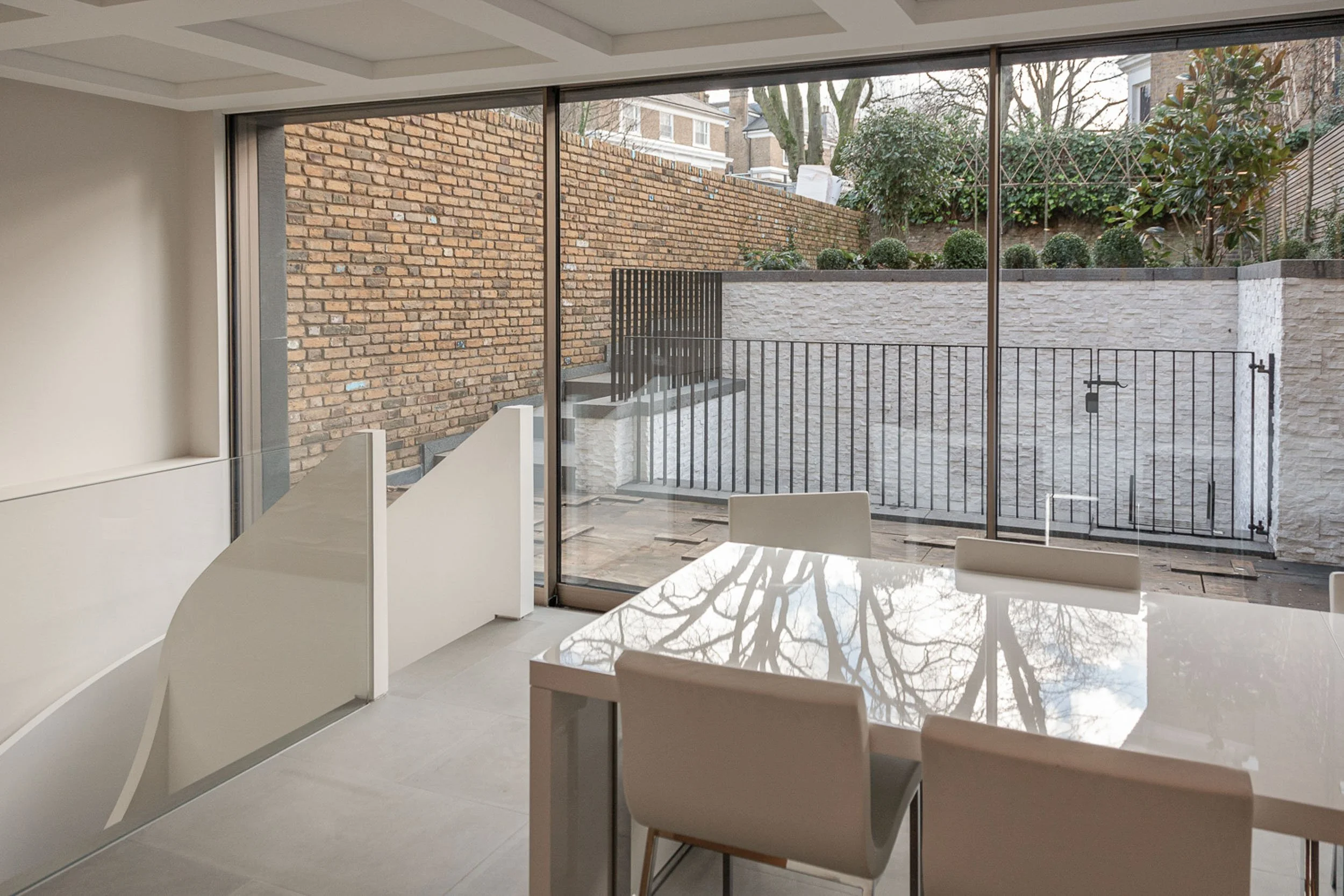 View from inside a modern room through large glass sliding doors to a backyard with brick walls, stairs, plants, and trees.