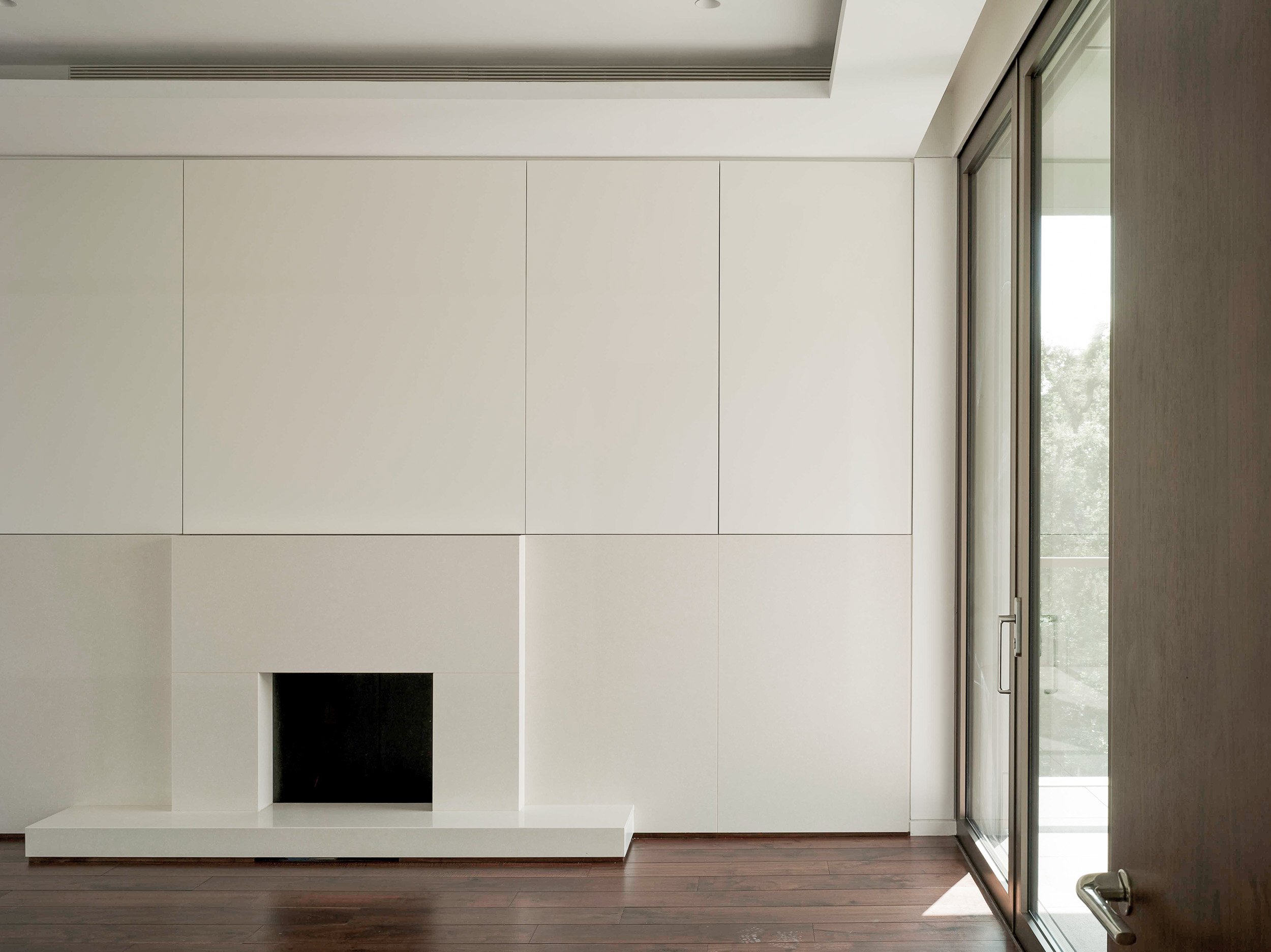 Contemporary indoor space with a white wall, a small black square opening, a wooden floor, and large glass sliding doors with visible trees outside.