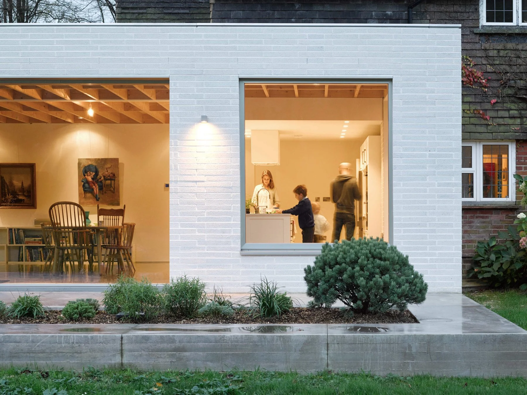 View of a modern house with a large window showing a family inside cooking in the kitchen and sitting at a dining table, outdoor garden with shrubs, and a concrete patio.