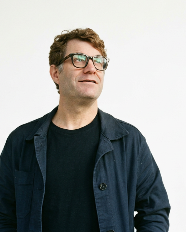 A man with brown curly hair wearing glasses, a black T-shirt, and a navy blue jacket posing against a white background.