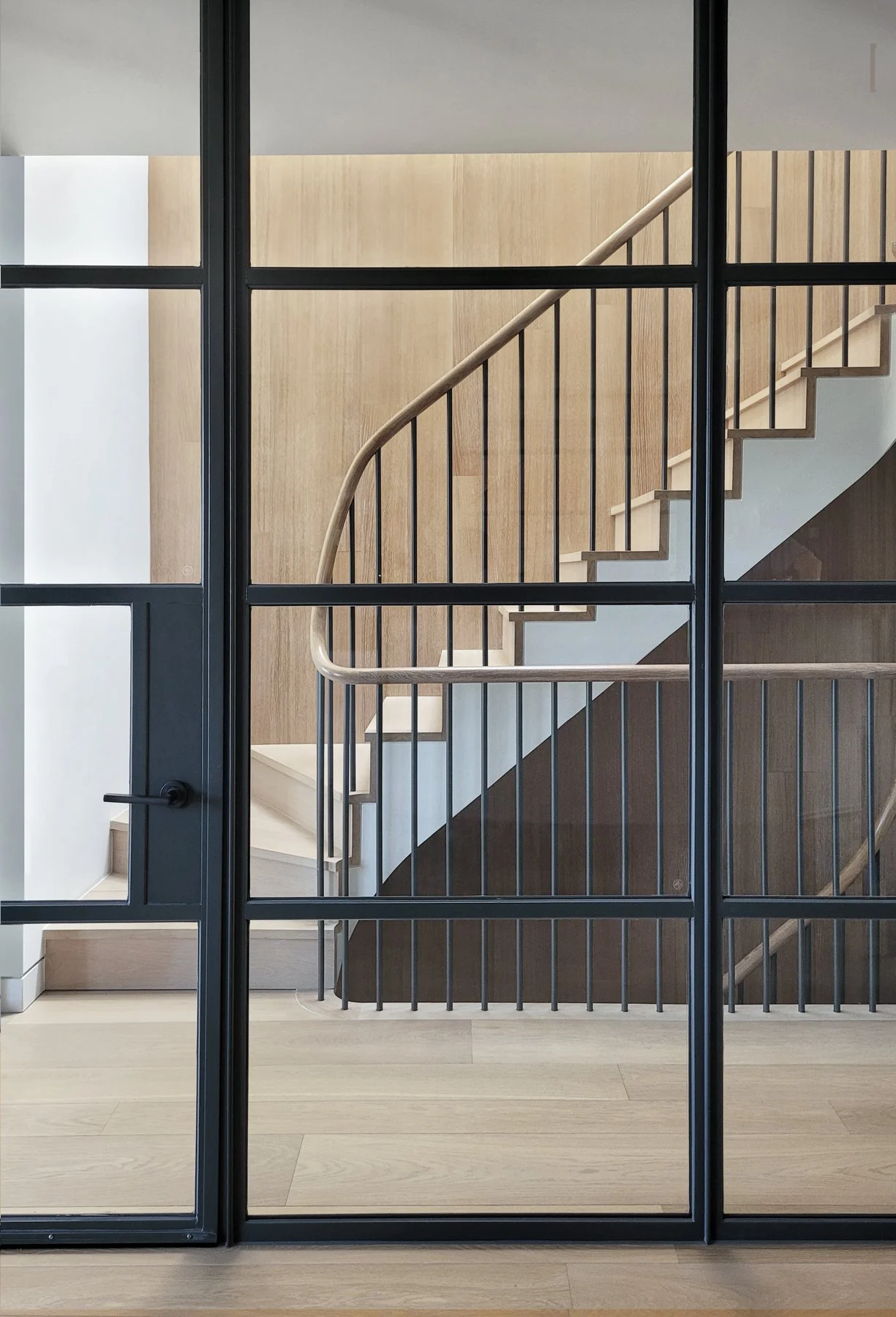 Interior staircase viewed through a black-framed glass door, featuring beige and dark brown wooden panels and a light-colored handrail.
