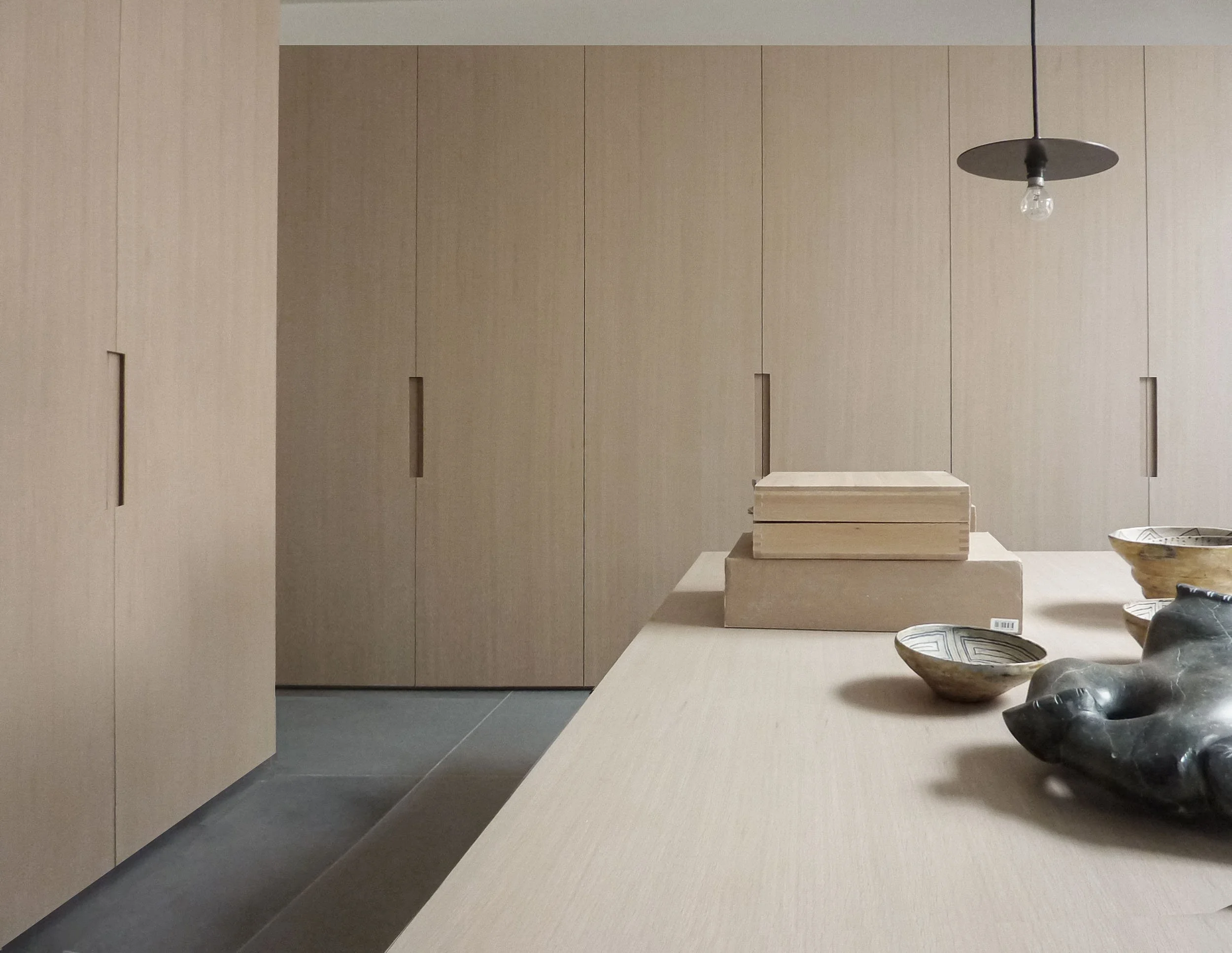 A modern interior with beige wooden panels on the walls, a hanging black lamp with an exposed light bulb, a stack of two beige boxes on a beige countertop, and various pottery bowls on the right side.