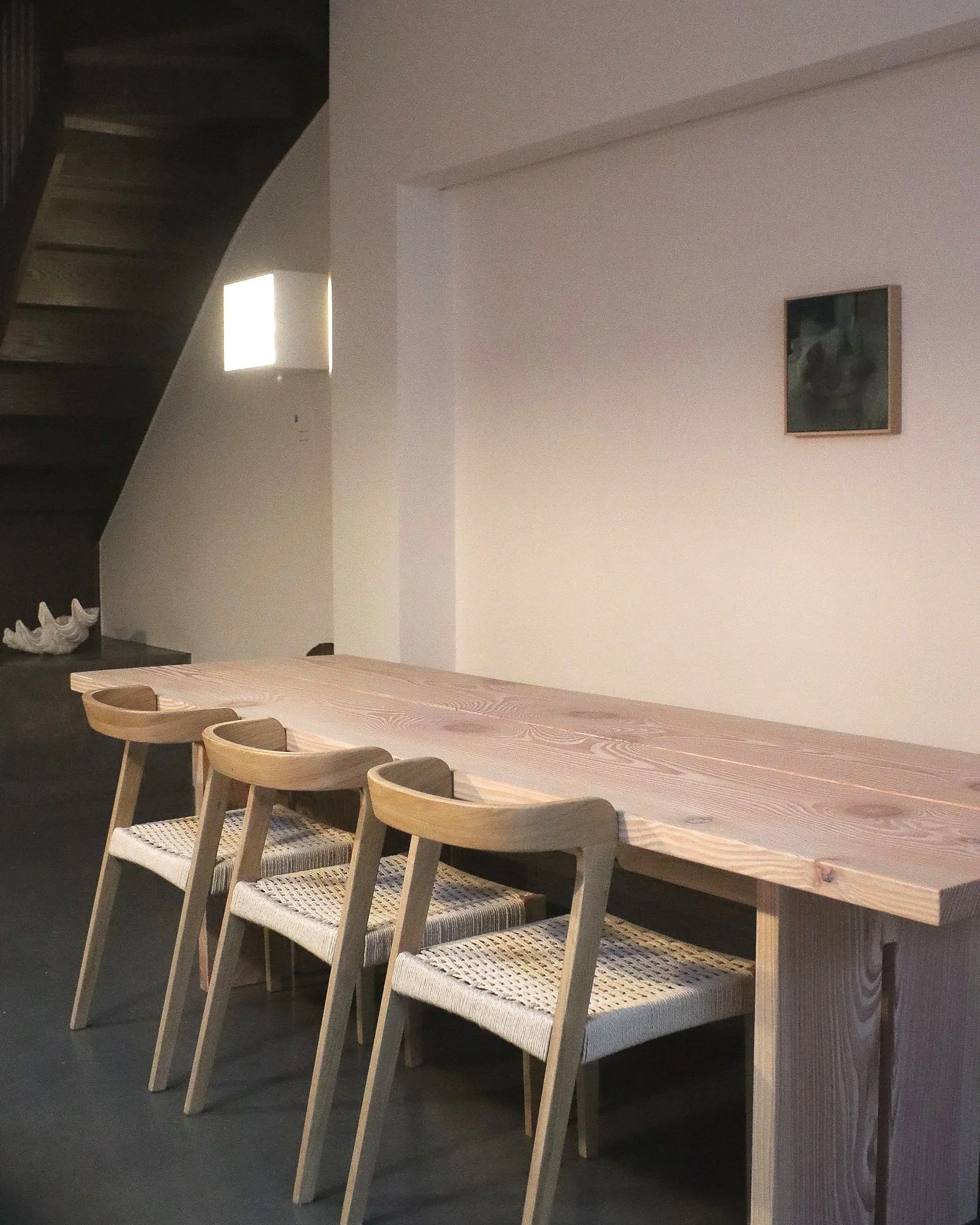 A modern dining area with a long wooden table and six chairs with woven seats. Behind the table, there is a white wall with a small framed picture and a staircase on the left. A small window is visible near the staircase, letting in light.