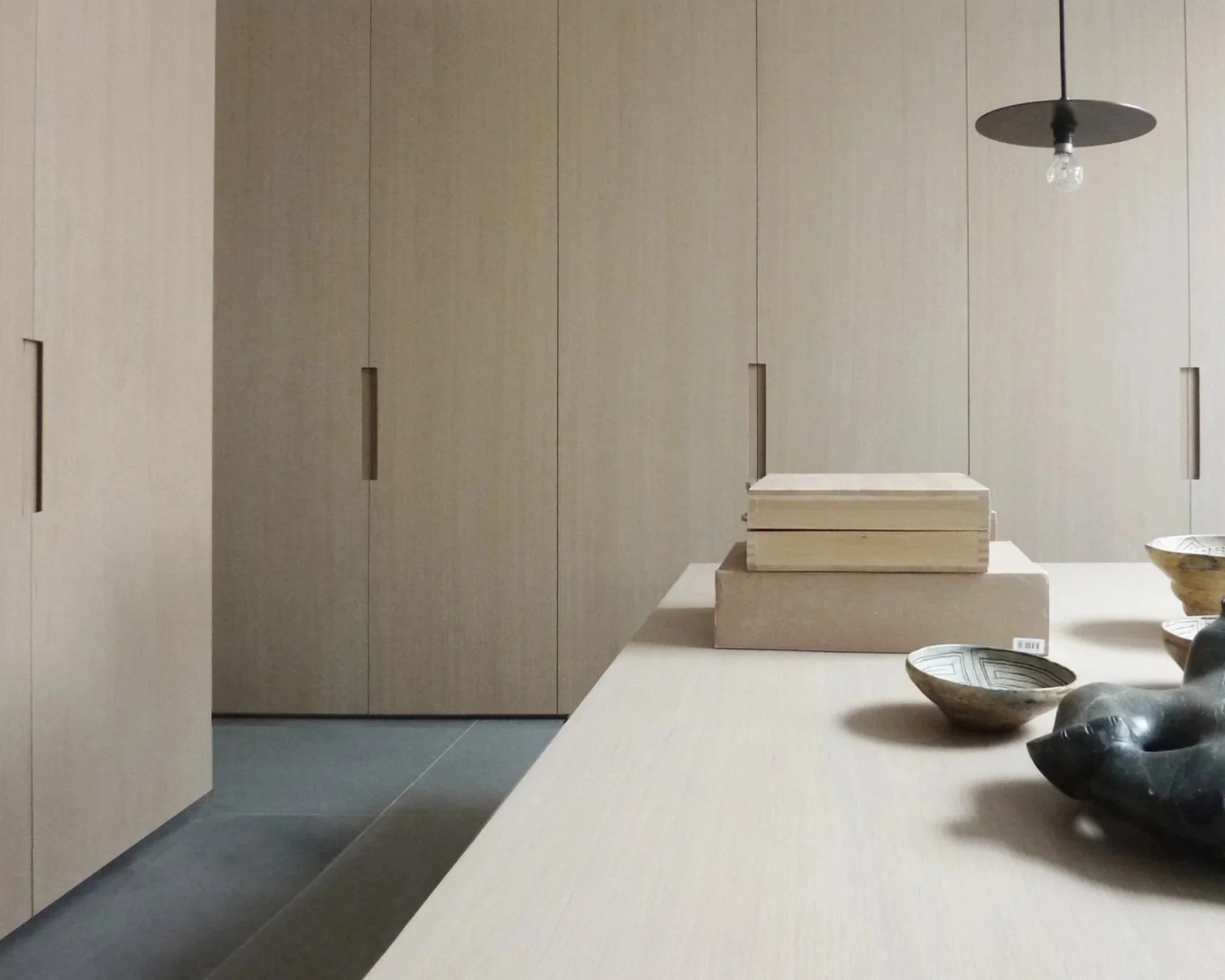 Minimalist interior with beige wall cabinets, a white table with wooden blocks, and ceramic bowls and stones on the table, illuminated by hanging pendant light with exposed bulb.