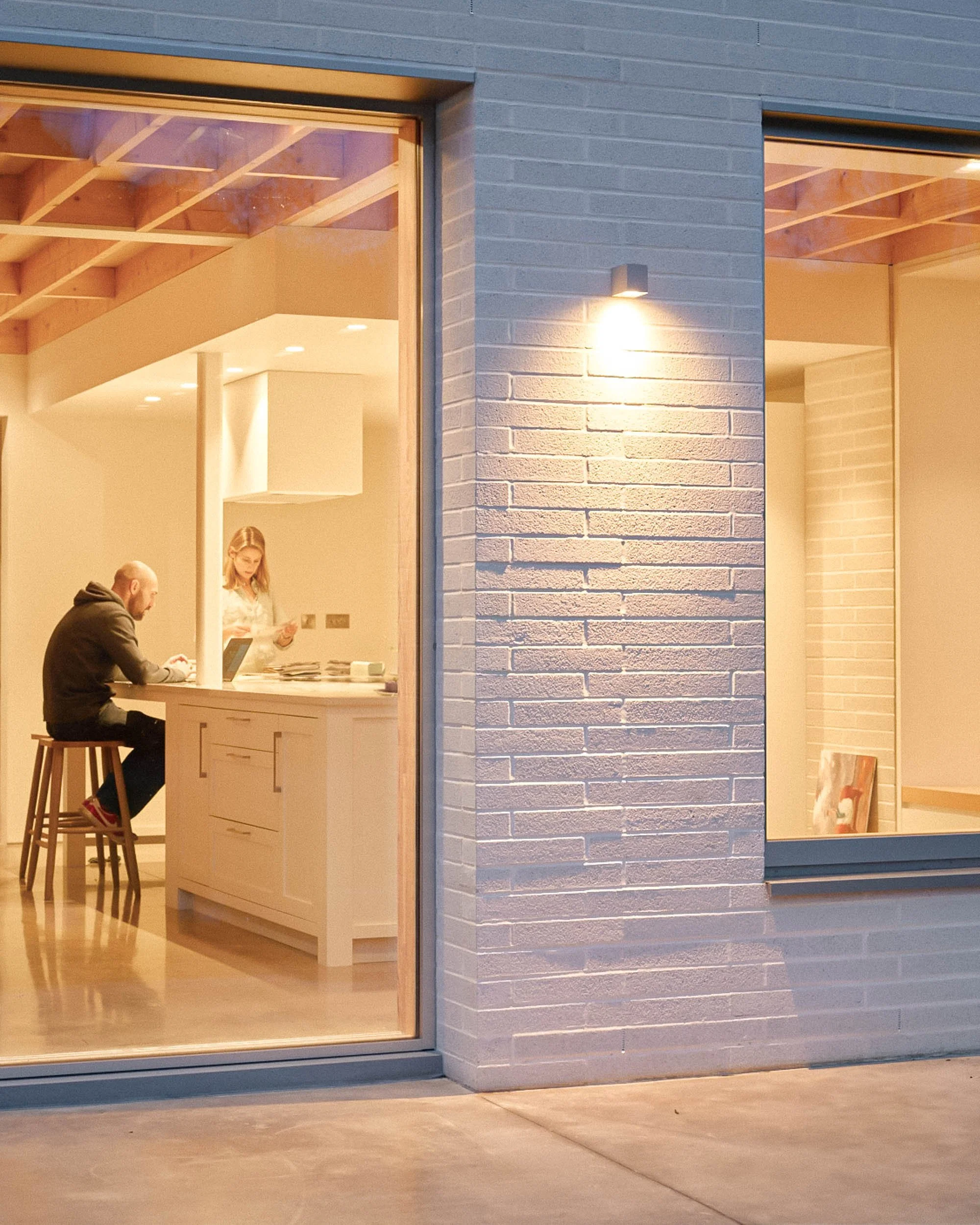 Inside view of a modern furniture shop with people sitting and standing at a counter, seen through a large window with brick walls outside and warm interior lighting.