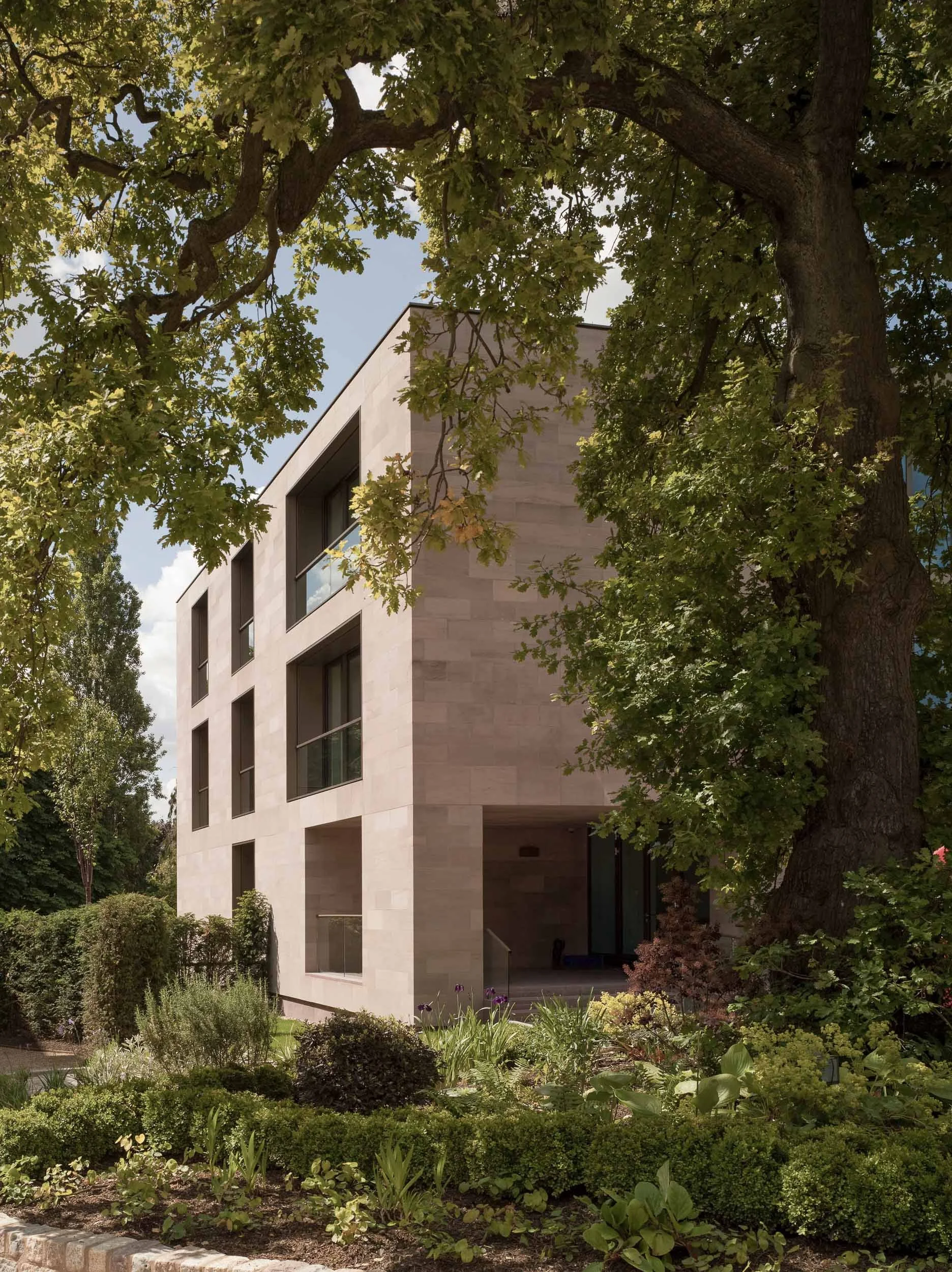 A modern, multi-story residential building with large windows, partially obscured by large, leafy trees, and surrounded by a garden with various plants and flowers.