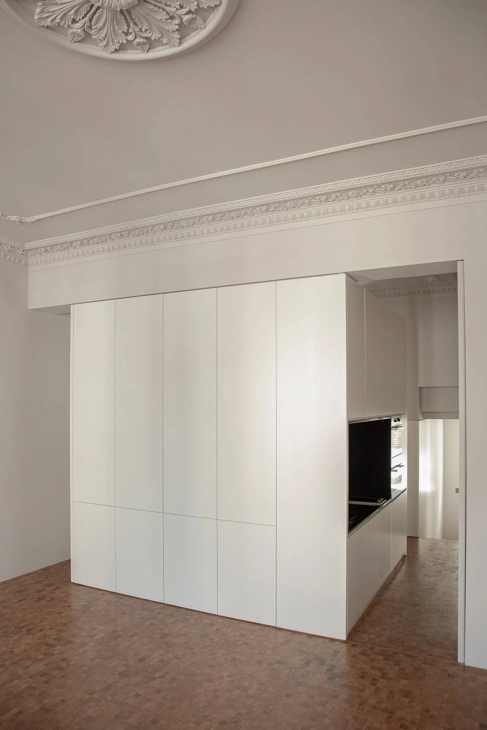 Modern white kitchen with cabinetry, built-in appliances, and a brown patterned wood floor, with decorative ceiling molding.