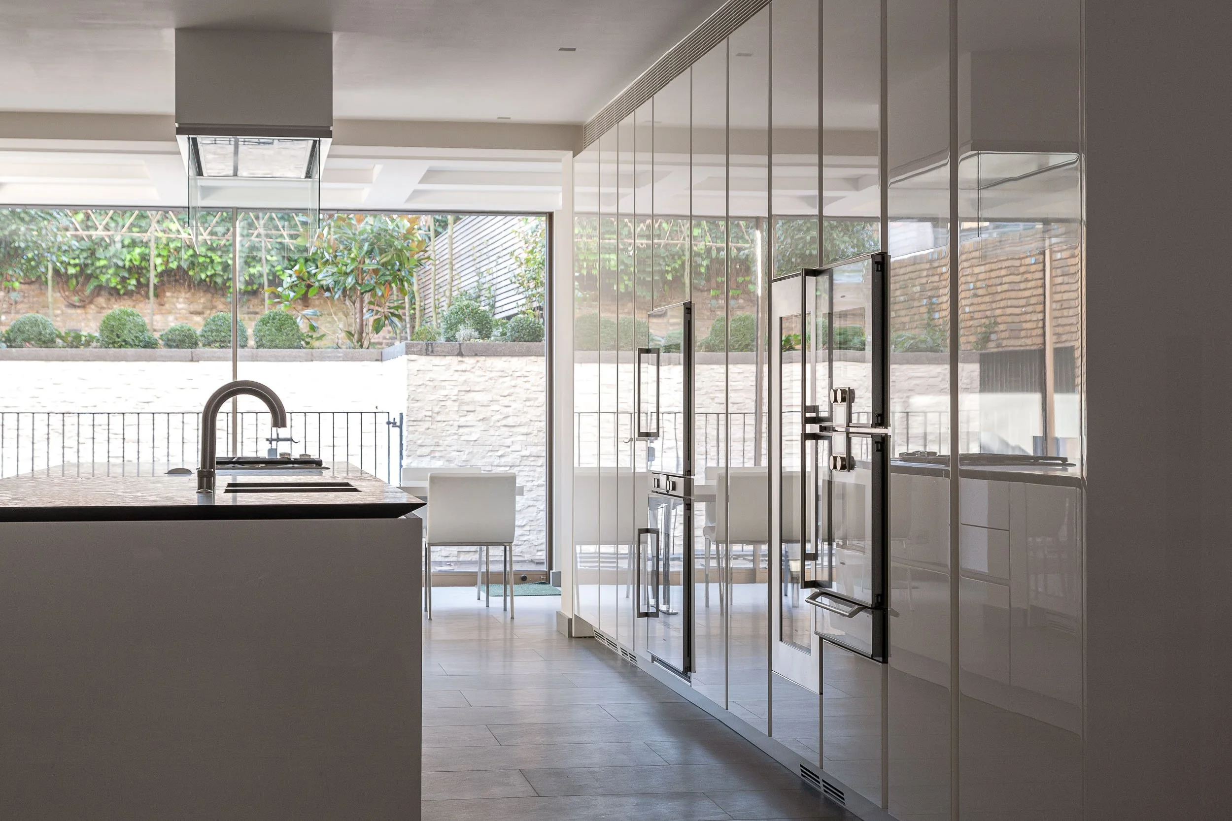 Modern kitchen with white cabinetry, glass doors, and a view of a backyard garden with trees and bushes.