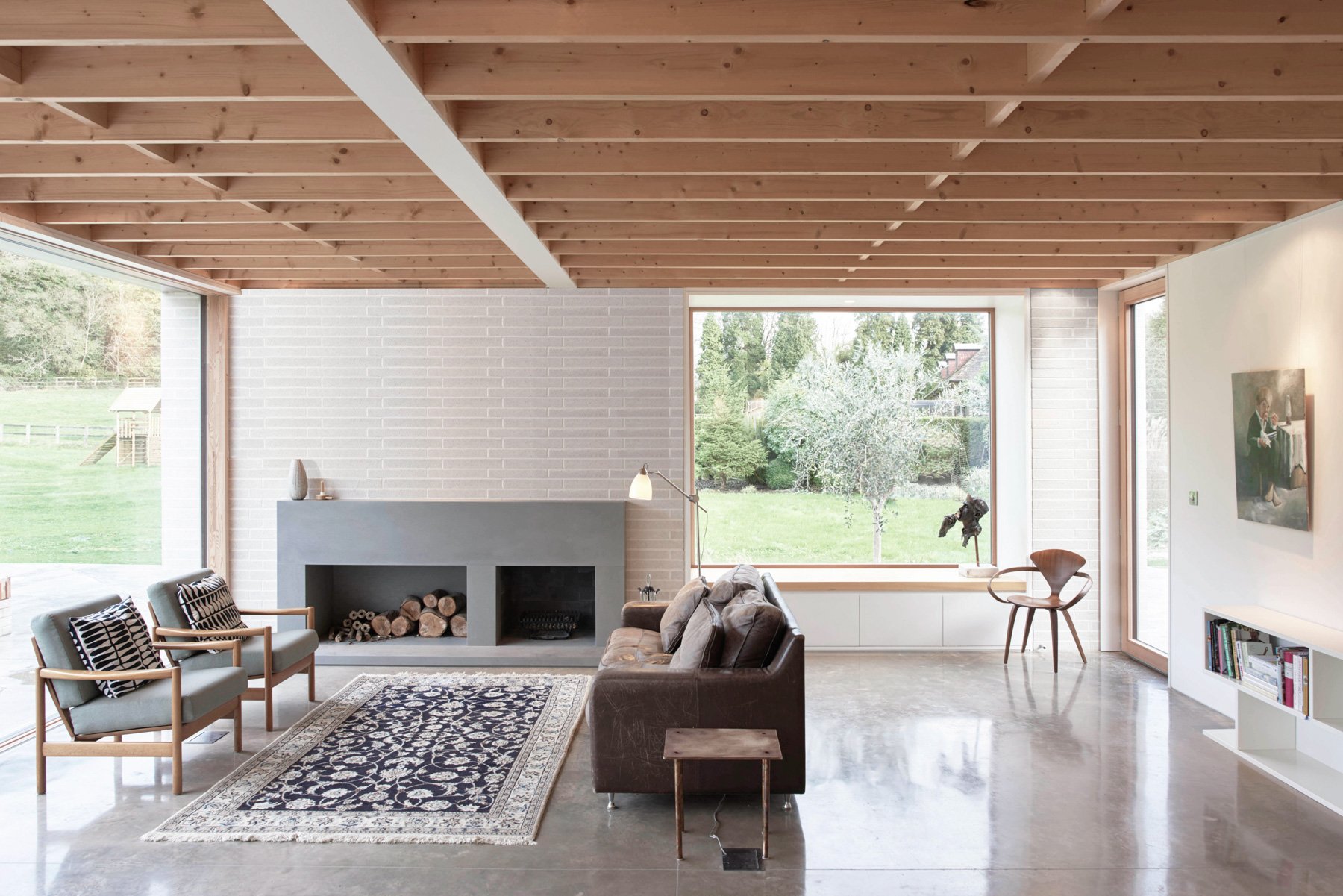 Living room with large windows overlooking a green yard, featuring a gray fireplace, a leather sofa, two armchairs, a patterned rug, a wooden side table, a floor lamp, and a bookshelf, with a wooden ceiling.