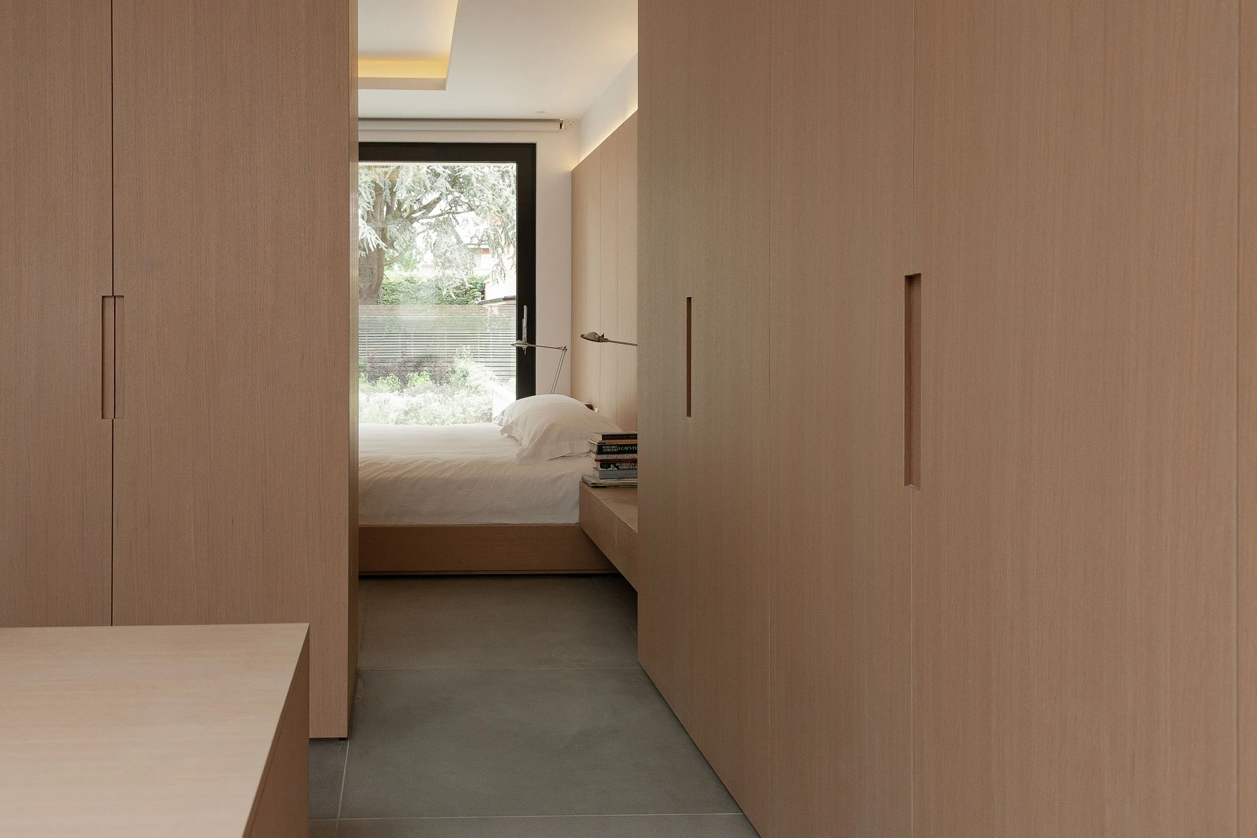 A minimalist bedroom with a large window showing greenery outside. The room has light wooden built-in cabinets and a bed with white linens. A small stack of books is on a bedside table.