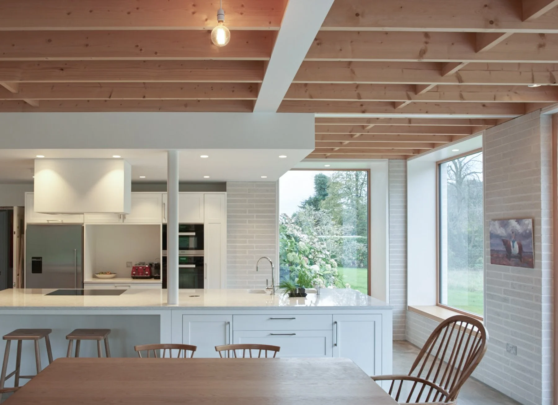 Modern kitchen with white cabinets, a large window showing greenery outside, and a wooden ceiling. There are four wooden chairs and a wooden dining table in the foreground.