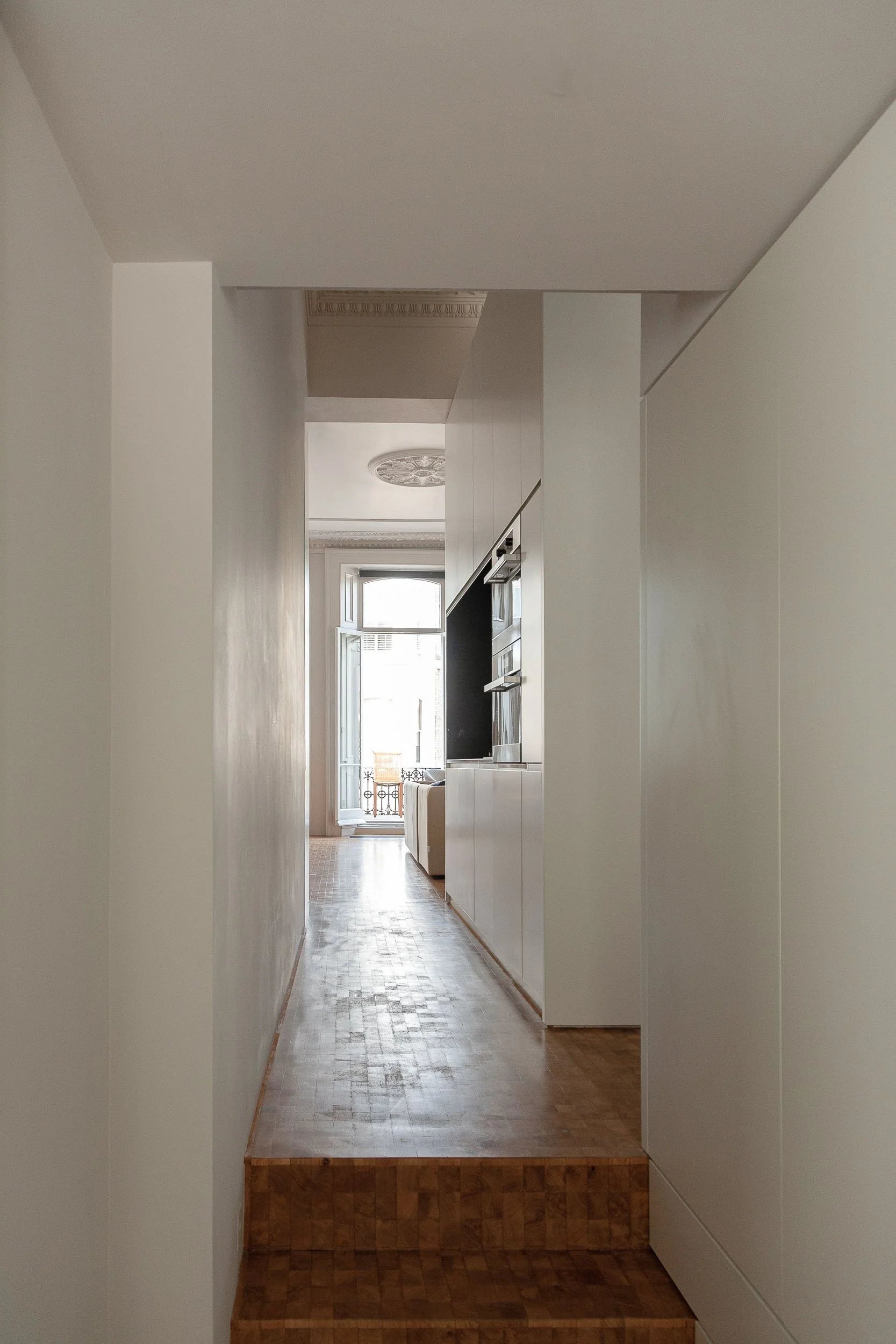 A narrow hallway extending toward a bright open window with a small balcony outside, visible in the background.