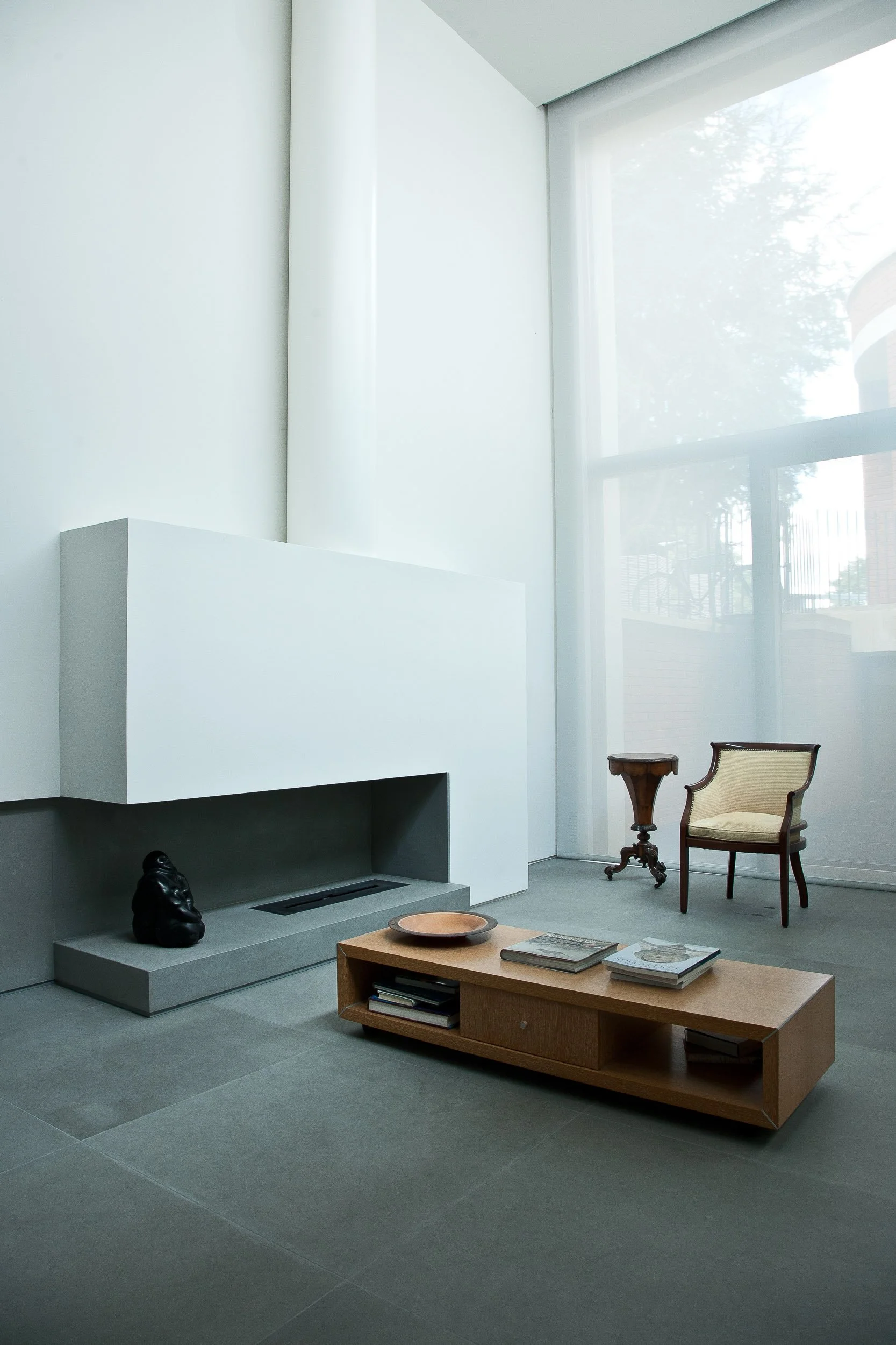 A modern, minimalistic living room with large windows, a white fireplace, a wooden coffee table with books and a bowl, a vintage-style armchair, a small wooden side table, and a black decorative sculpture near the fireplace.