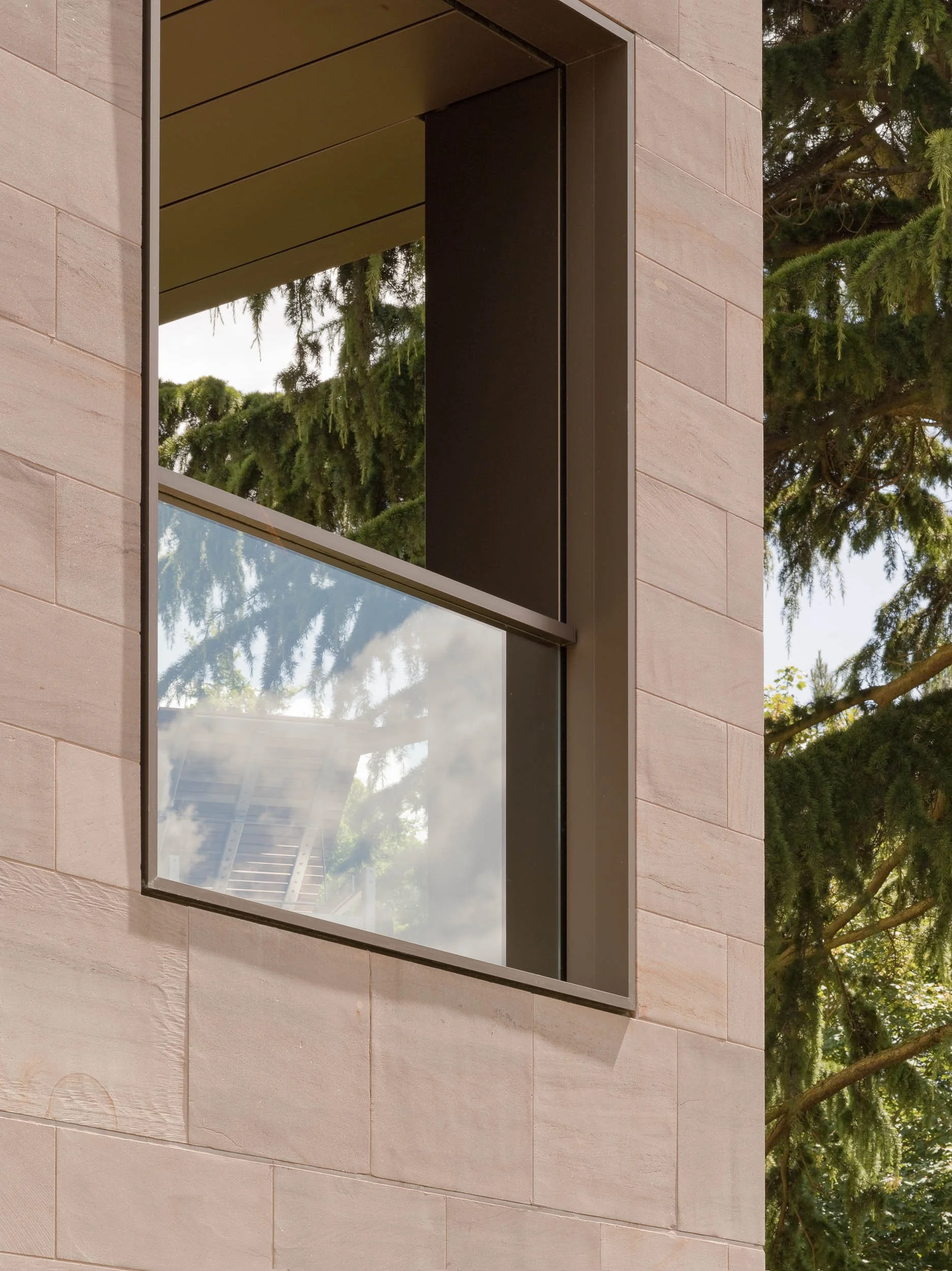 Close-up of a window on a modern building with beige stone exterior walls, reflecting trees and sky.