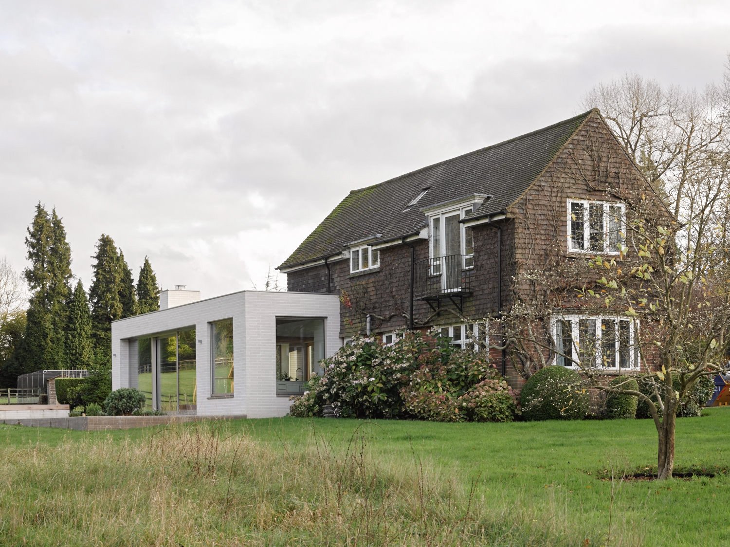 Traditional brick house with brown shingles and white-framed windows, alongside a modern white extension with large glass windows, surrounded by a well-maintained lawn and flowering bushes.