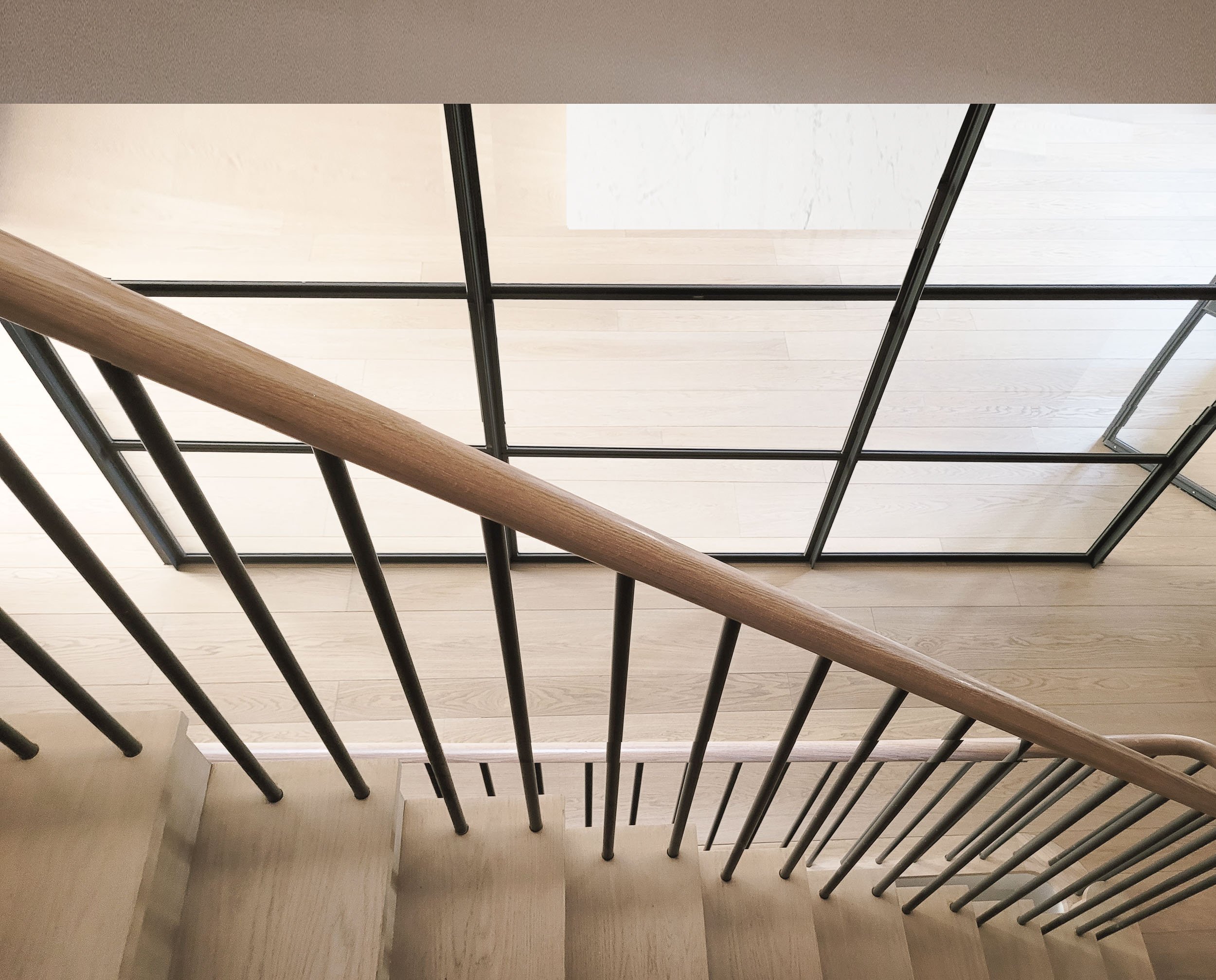 Downward view of a staircase with black metal railing and wooden handrail, looking at a light-colored wooden floor and a white wall with a window.
