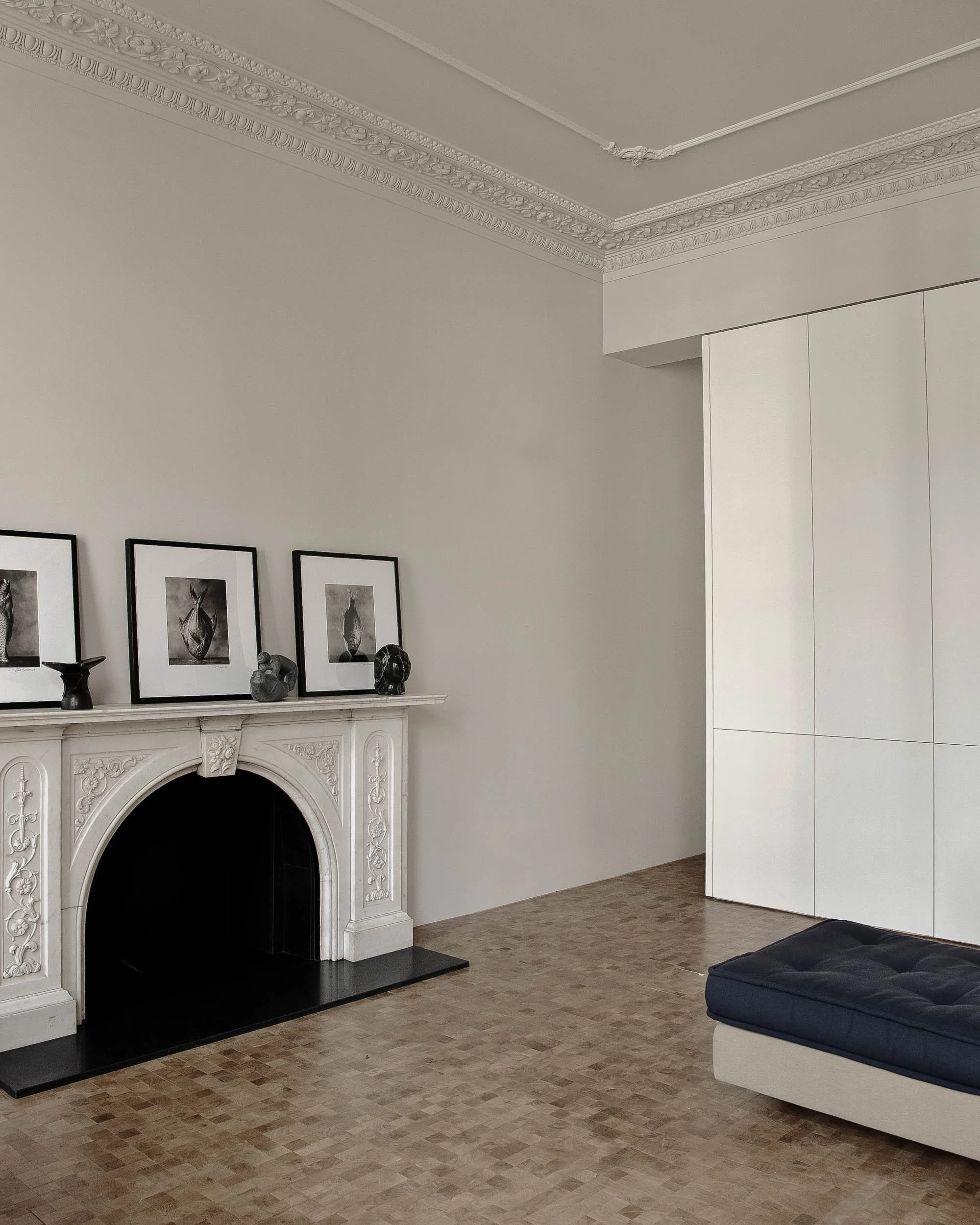 A living room with a white ornate fireplace, black interior, and decorative objects including framed art, sculptures, and a candlestick on the mantel. Light-colored wood flooring, a part of a dark blue sofa on the right, and a white modern cabinet or partition are visible.