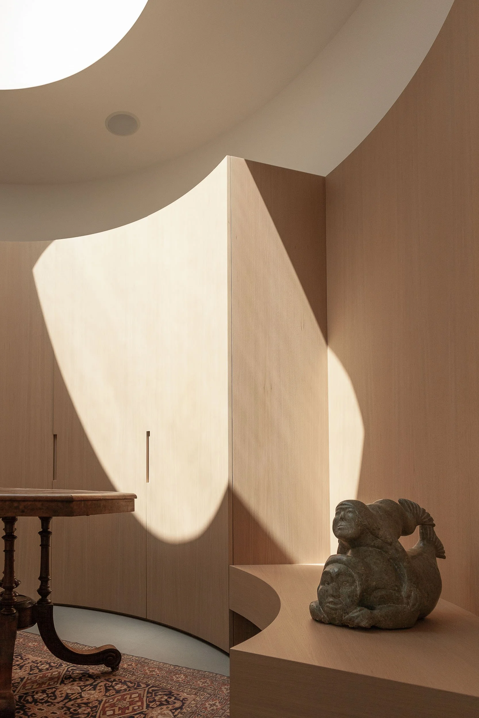 A modern interior with a beige curved wall, a carved sculpture of a reclining figure, a wooden table with chairs, and patterned rug on the floor. Natural light creates shadows on the wall.