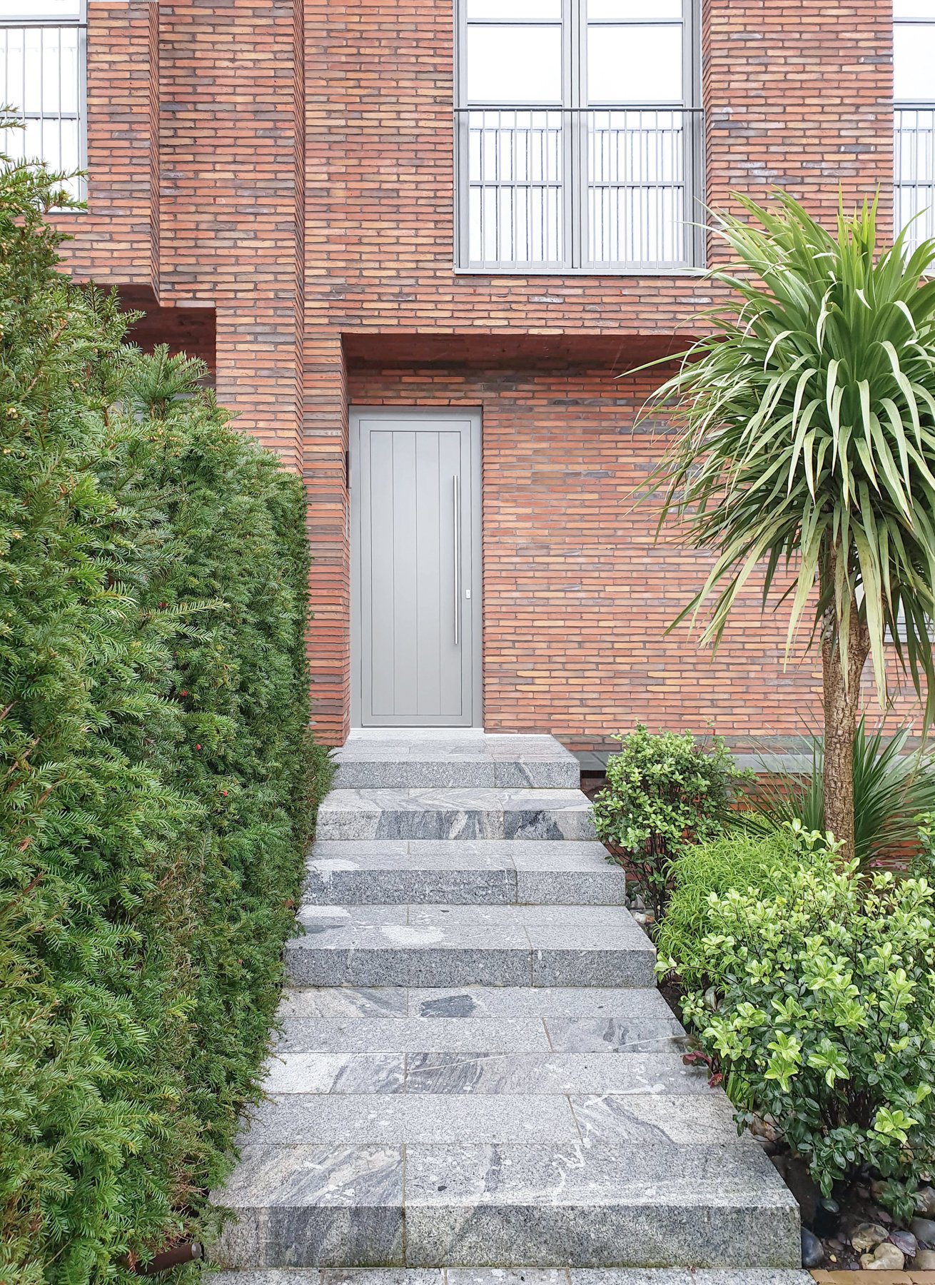 A modern brick house entrance with a gray door, gray stone steps, green bushes and a tall tree on the right side.