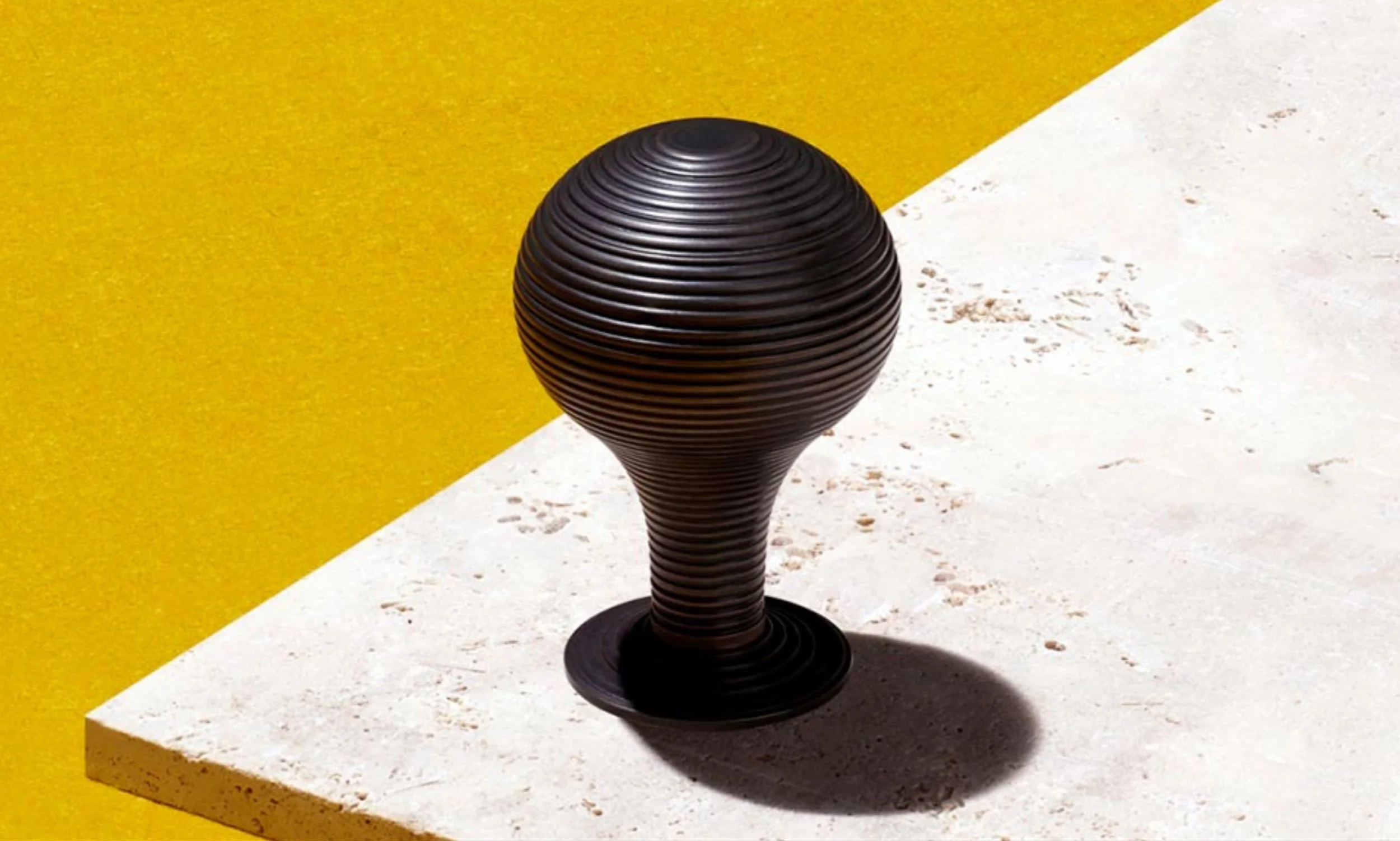 Black modern decorative sculpture with horizontal ridges, placed on a stone pedestal against yellow and white background.