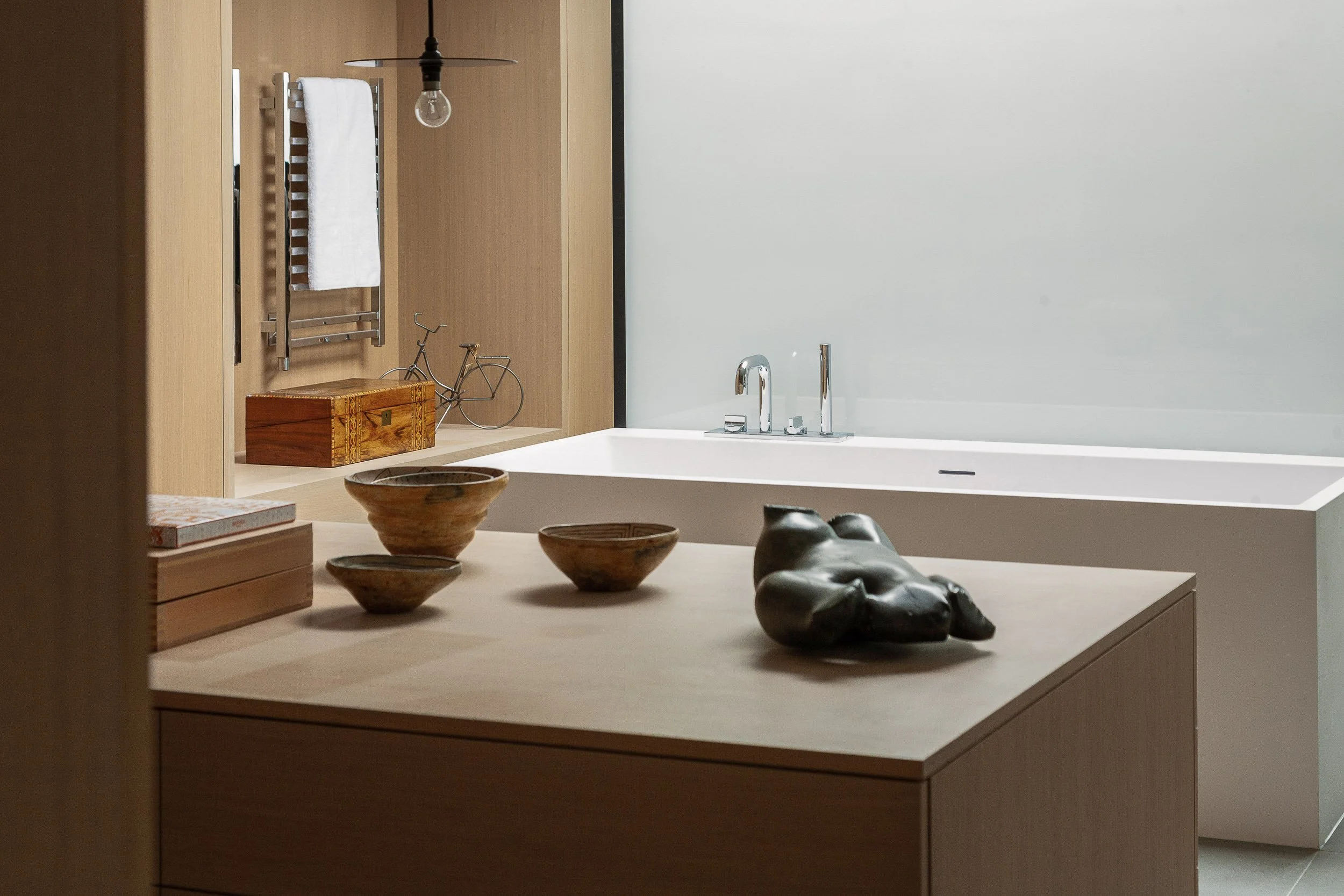 A minimalist bathroom with a wooden wall and a white bathtub. On the countertop, there are decorative bowls, stacked books, and a small wooden box. In the background, a wall-mounted towel rack with a white towel, a bicycle sculpture, and a wooden box are visible. A hanging light bulb is also seen.