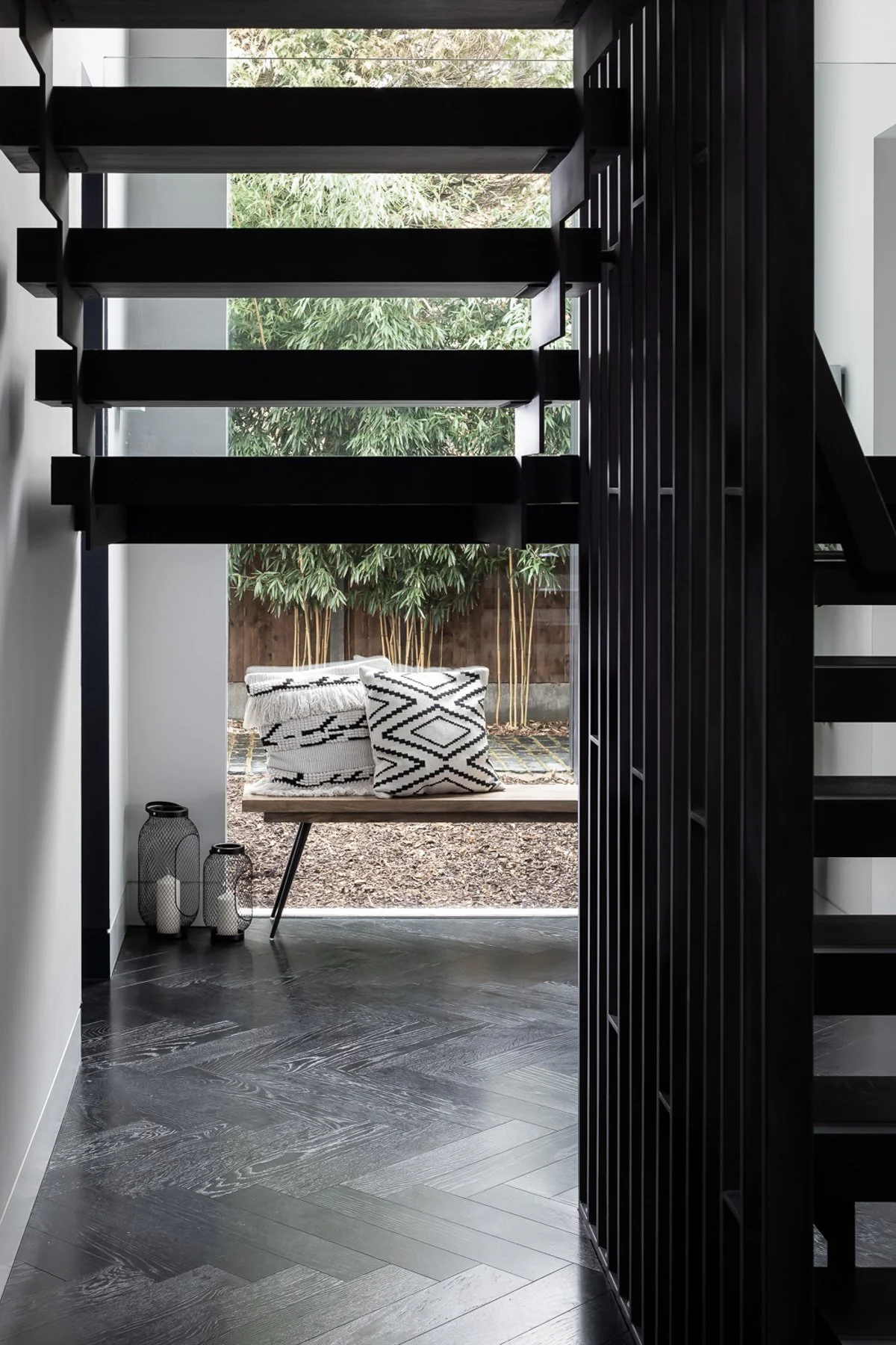 Interior view of a modern black staircase with open risers, leading to an upper level, adjacent to a glass wall overlooking a backyard with bamboo plants, featuring a wooden bench with black and white patterned pillows and decorative vases on the floor.