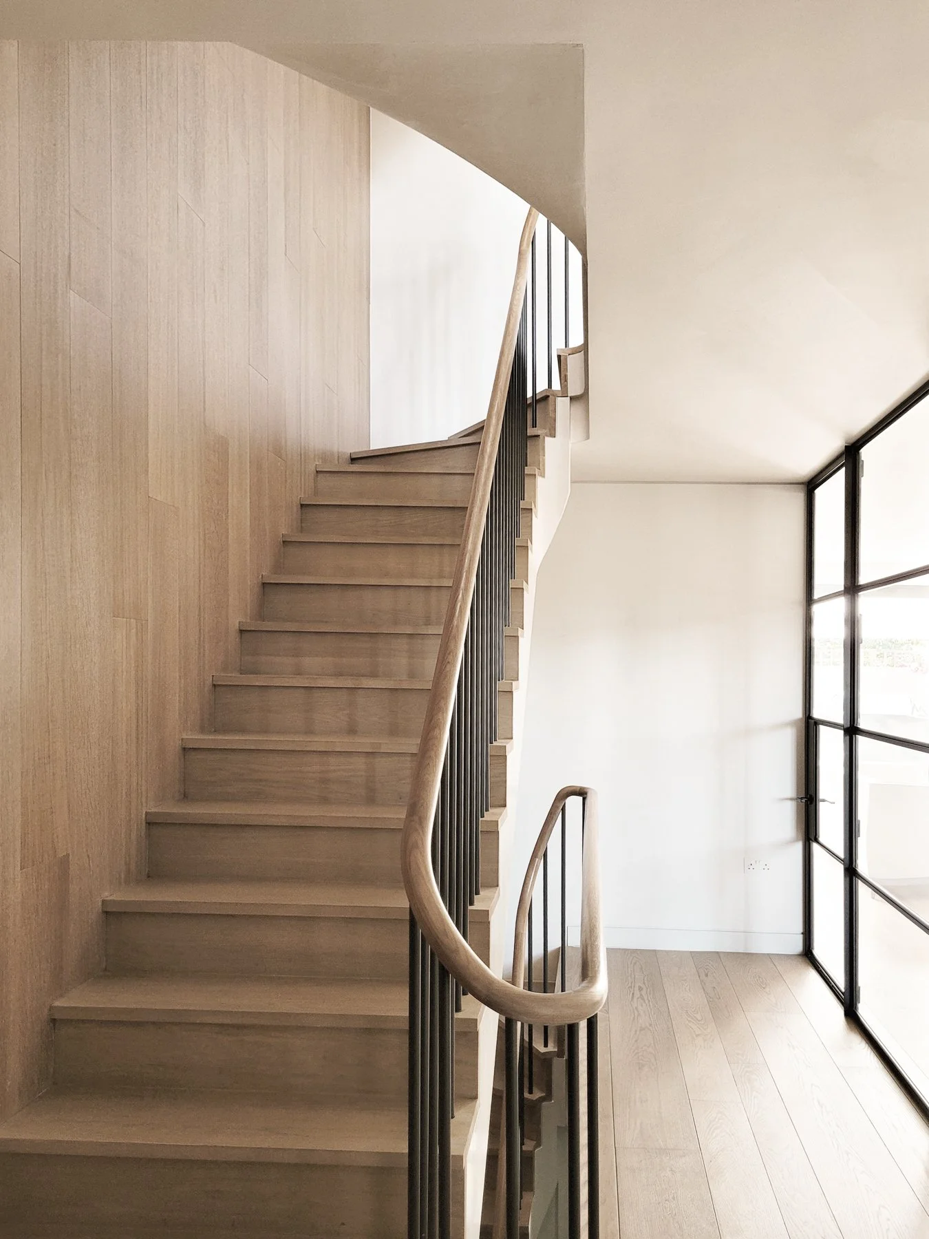 Interior wooden staircase with black railing and glass partition by large window in a modern minimalist home.