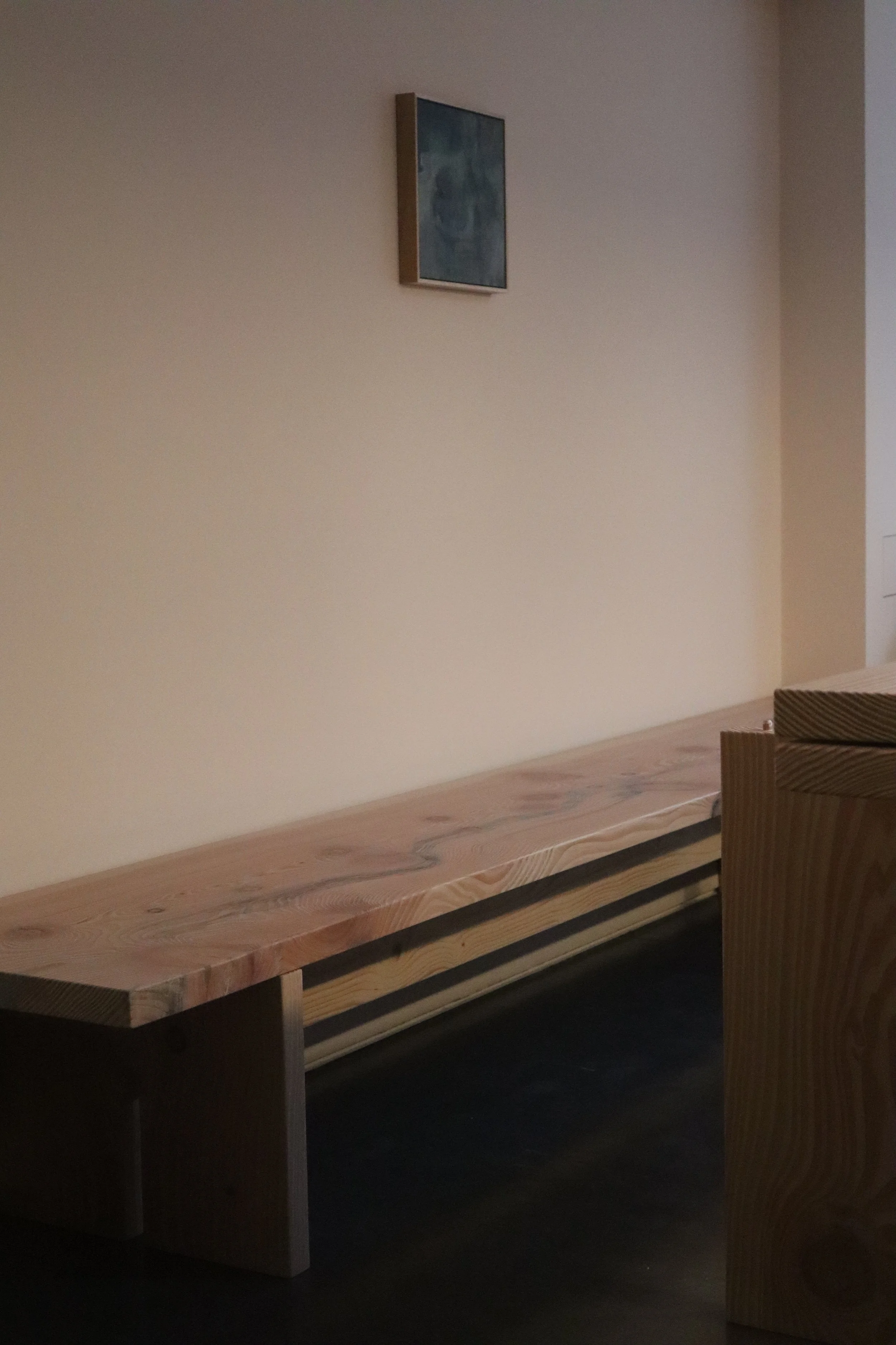 A long wooden counter or bench with a natural wood finish is placed against a plain beige wall. A framed abstract blue and green artwork hangs above. To the right, a portion of striped upholstered furniture is visible.