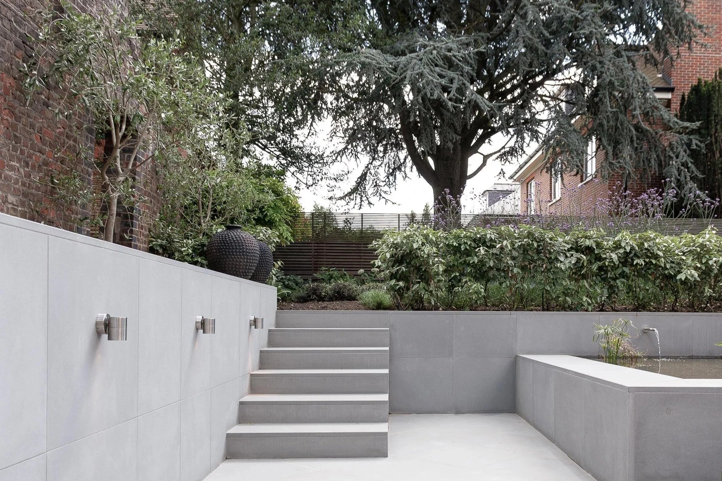 Modern outdoor patio area with light gray tiled walls and stairs, decorative black ceramic vases, lush green plants, a large tree, and a residential building with brick walls and windows in the background.