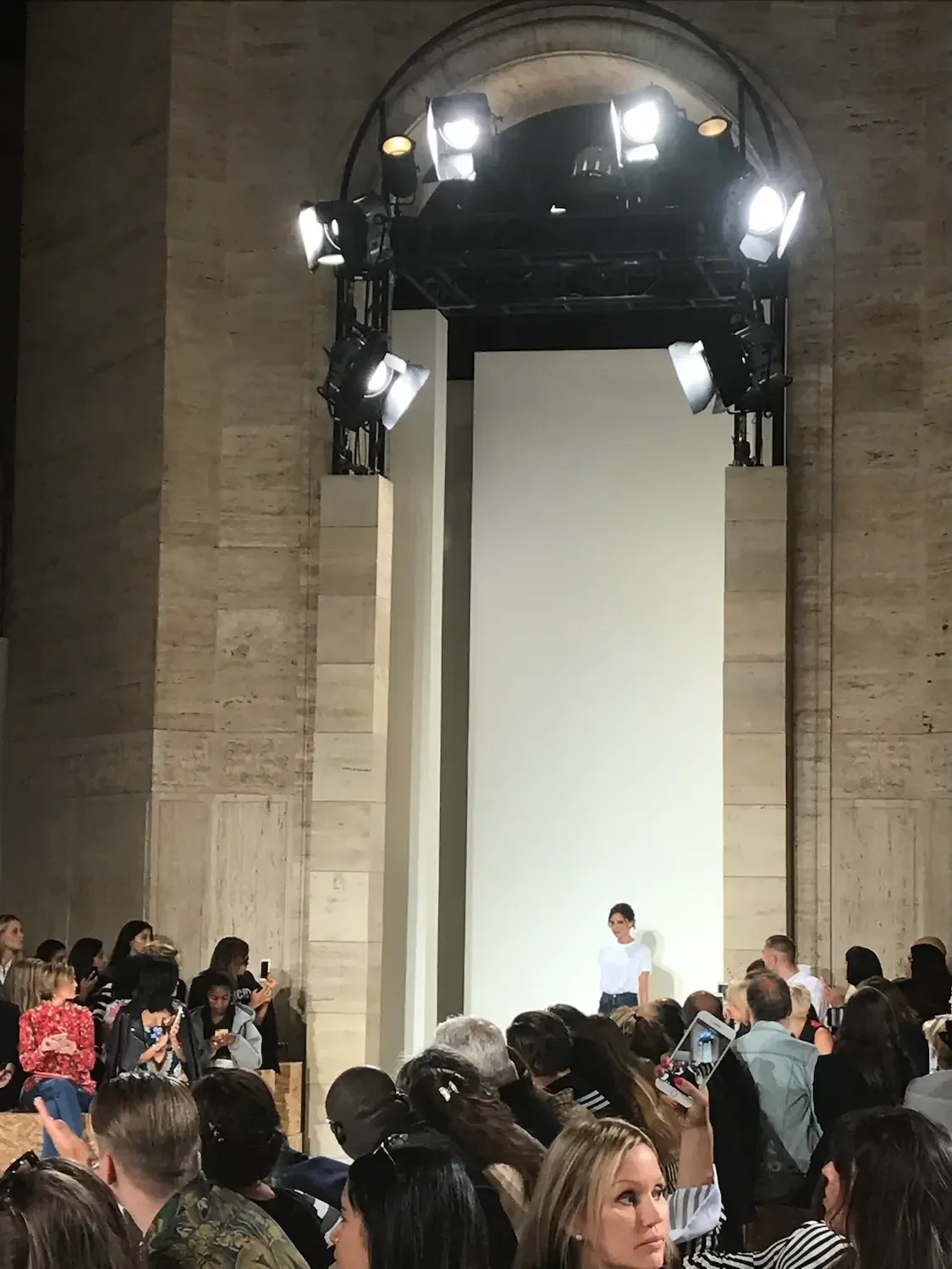 Fashion show runway with model standing on stage, audience seated around, bright lights overhead, and a large white backdrop behind the model.