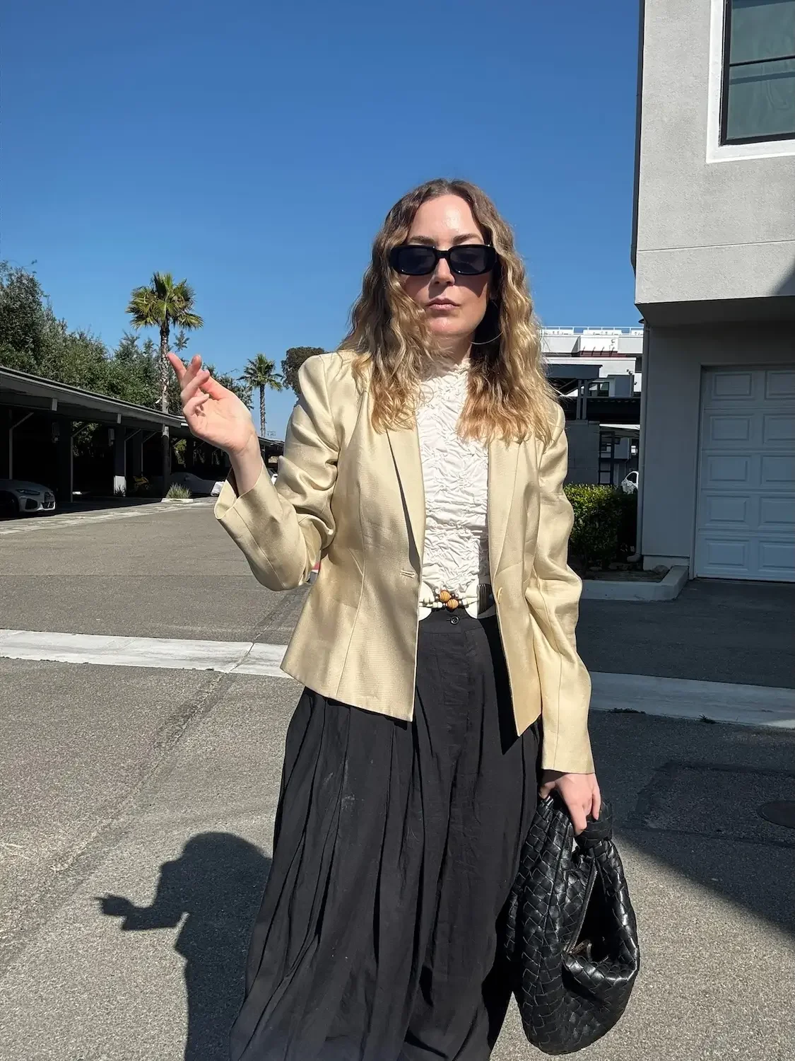 A woman with curly blonde hair wearing sunglasses, a cream blazer, and black wide-leg pants standing outdoors on a sunny day, holding a black woven handbag.