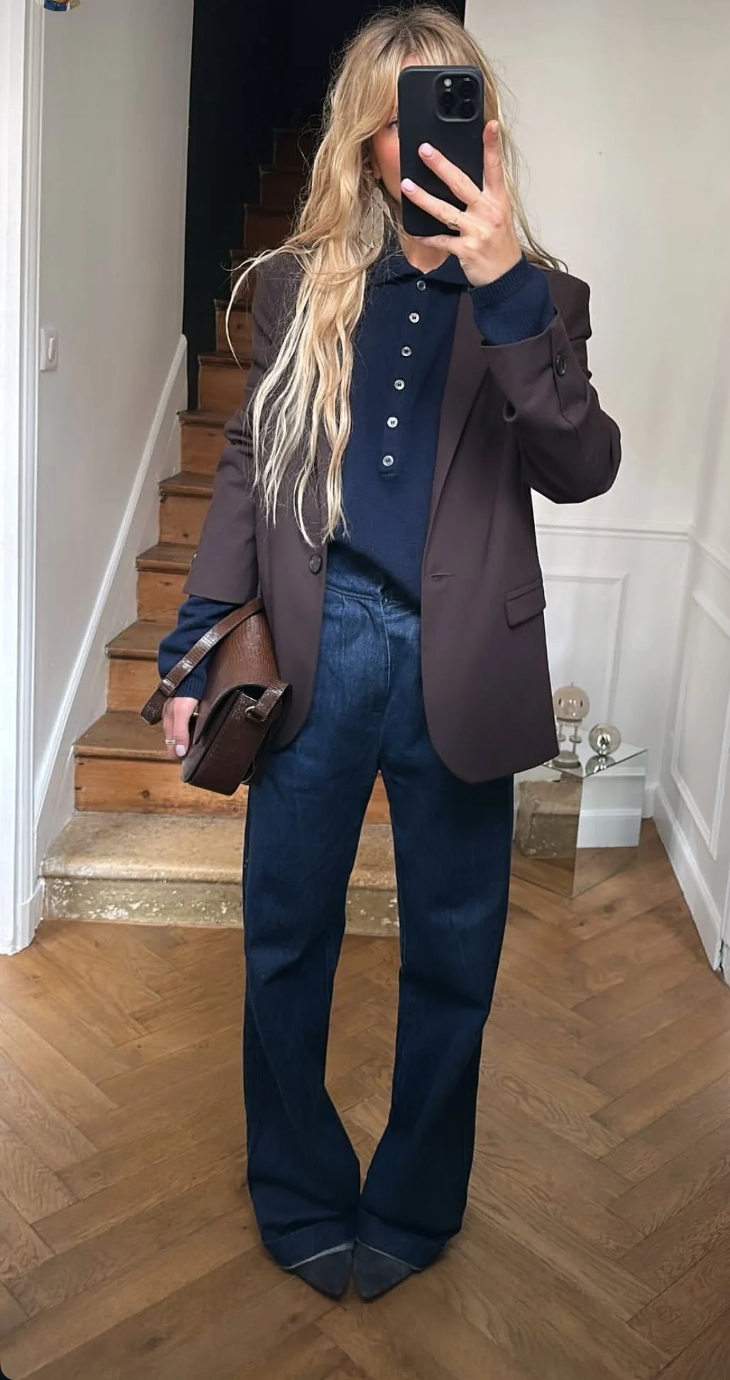Woman taking a mirror selfie indoors, wearing a brown blazer over a navy blue long-sleeve shirt, dark blue jeans, and gray pointed shoes, holding a brown purse.
