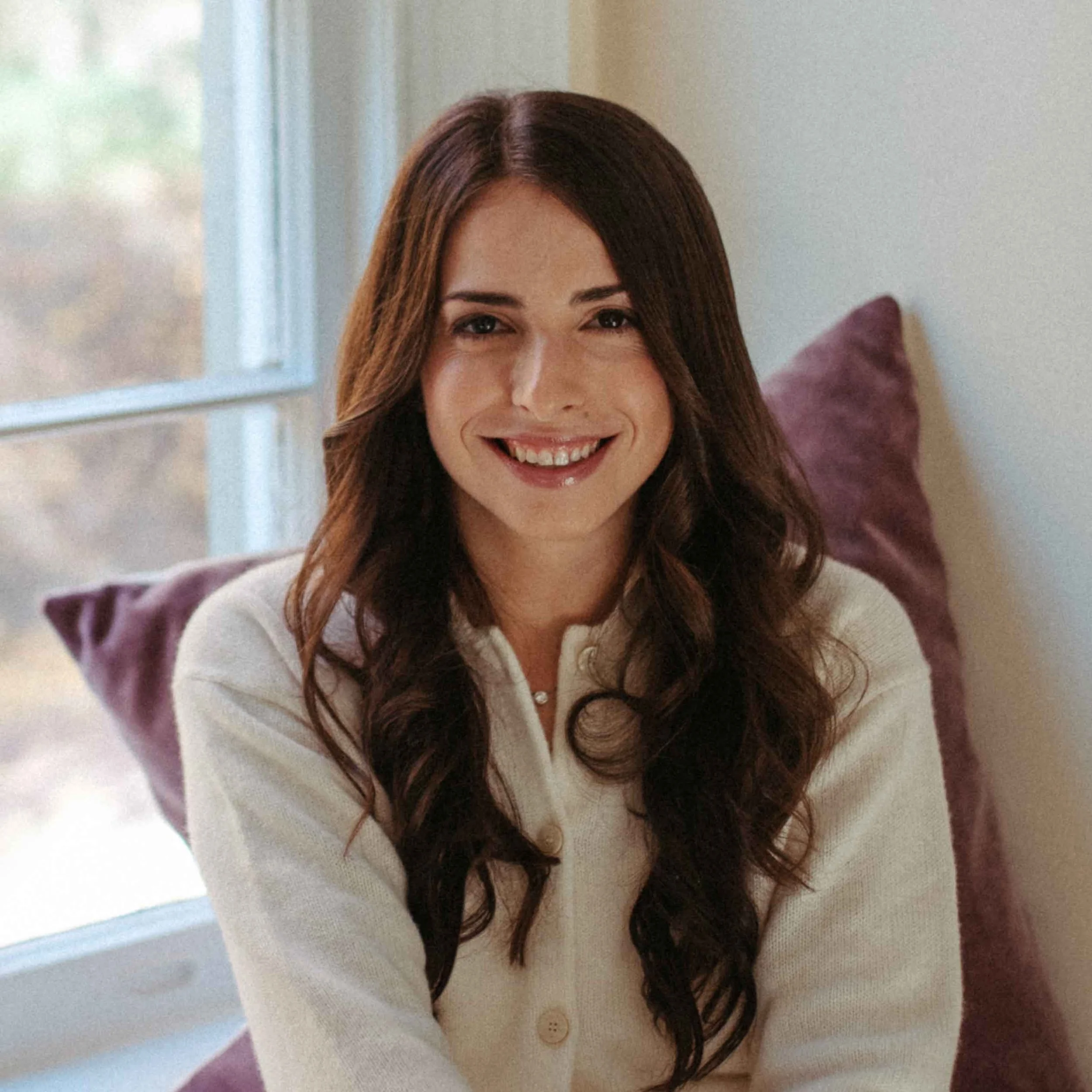 A woman with long brown curly hair smiling, sitting on a purple pillow near a window.