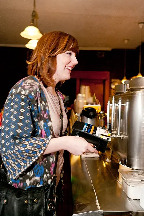 A woman with shoulder-length brown hair, wearing a colorful patterned jacket, is smiling while standing at a beverage counter holding a payment terminal.
