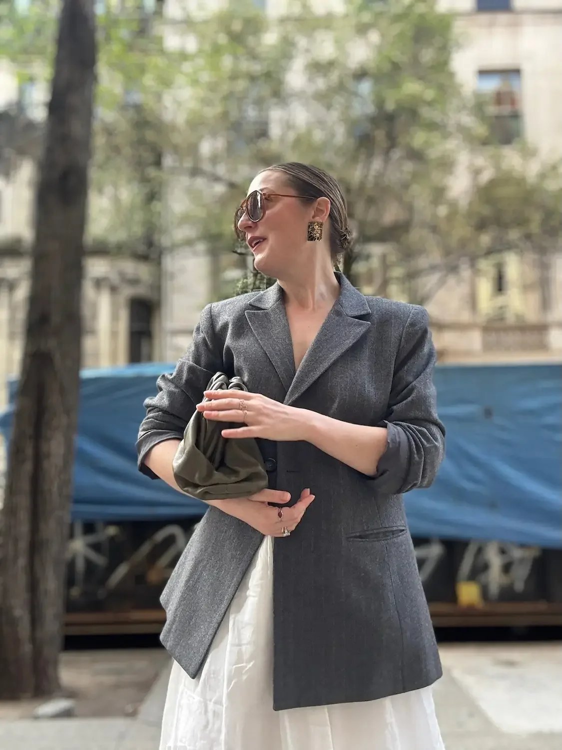 A woman with dark hair, wearing sunglasses, a gray blazer, and earrings, standing outdoors in front of a blurred background of trees and buildings, holding a small purse.