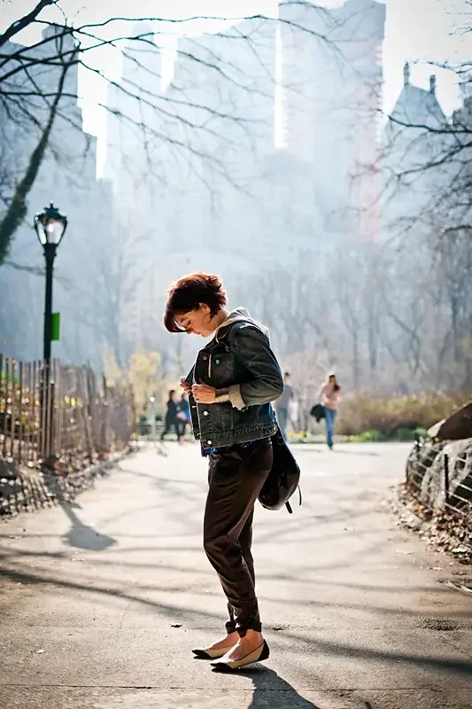 A woman in a denim jacket and black pants walking in a park on a sunny day, holding a phone, with other people in the background and tall buildings in the distance.