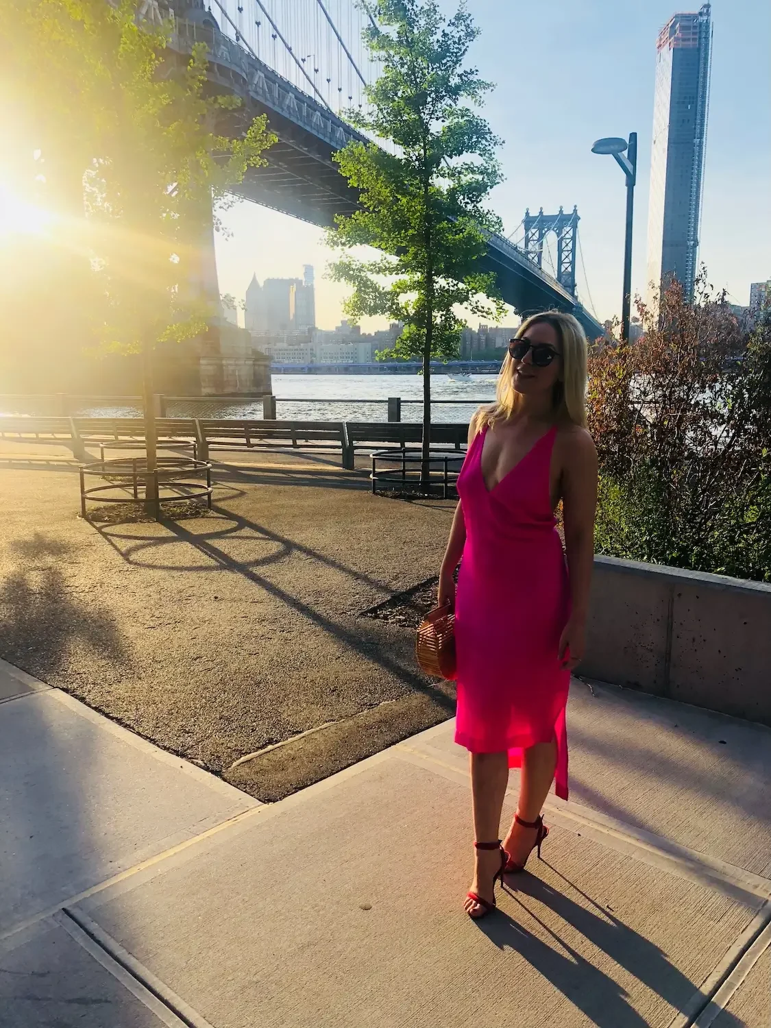 A woman in a bright pink dress and black sunglasses standing on a sidewalk near the water in New York City. The Brooklyn Bridge and city skyline are visible in the background during sunset.