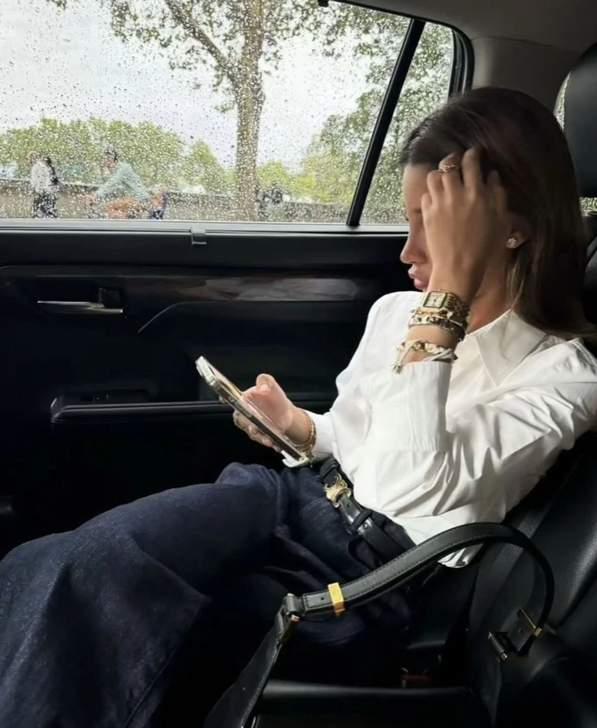 A woman in business attire sitting in a car, looking at her phone, with raindrops on the window outside.