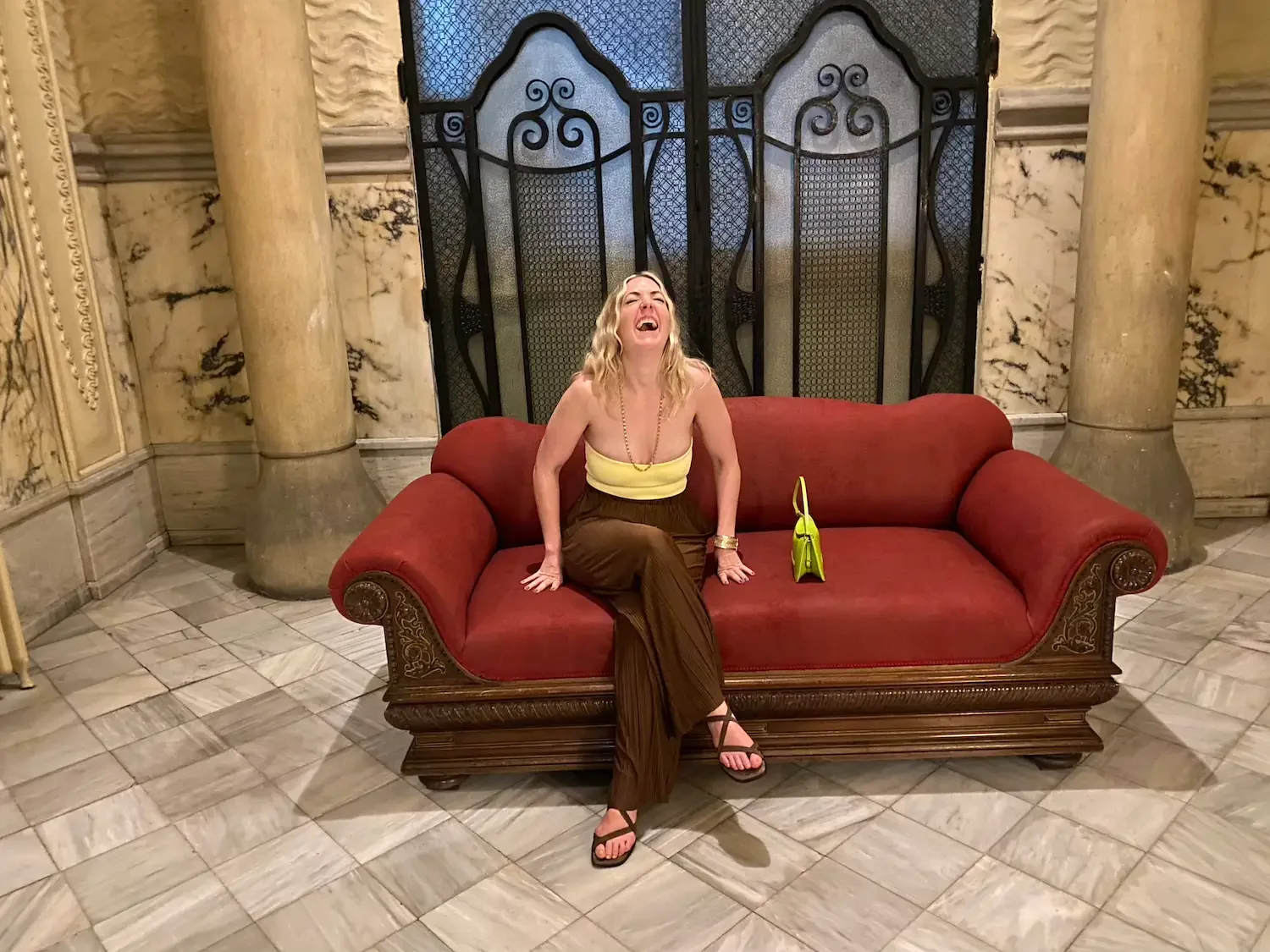 Nicole Russo wearing a yellow top and brown pants, sitting on a red velvet sofa, laughing. There is a yellow bag beside her on the sofa, and the background features ornate columns and decorative black wrought iron panels.