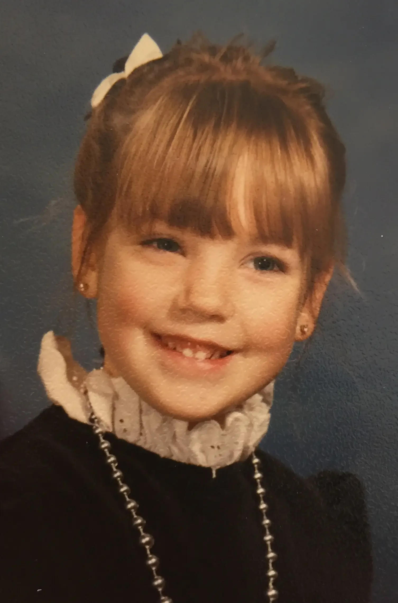 A young girl with light brown hair in a bun with a white bow, smiling with missing front teeth, wearing a black top, a white ruffled collar, and a pearl necklace, against a plain dark background.