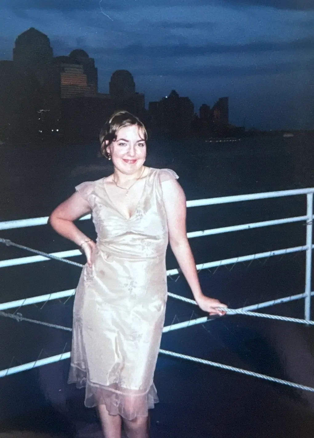 A woman in a beige satin dress standing on a boat deck at night with city skyline in the background.