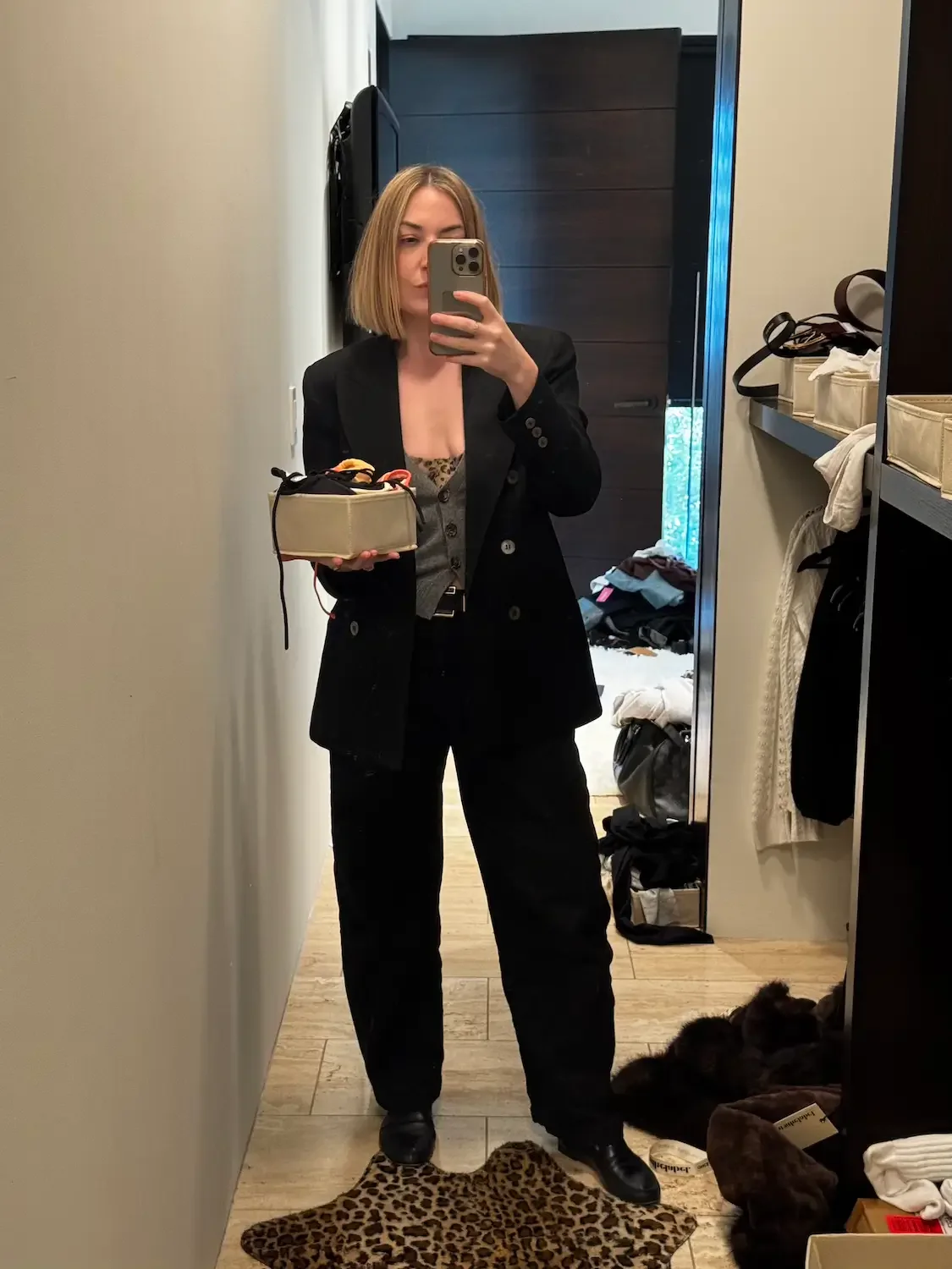 Nicole Russo taking a mirror selfie in a room with clothes and shoes on the floor. She is wearing a black blazer, black pants, and black shoes, holding a beige bag with some items inside.