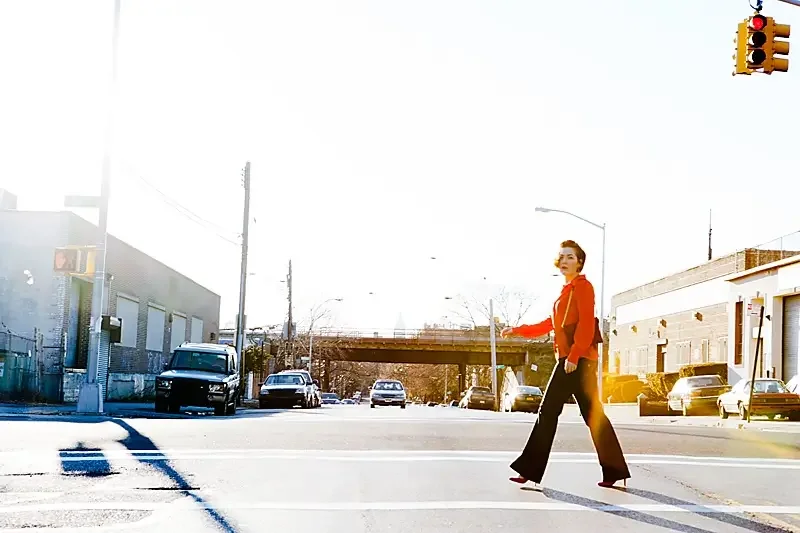 A woman crossing a city street at a crosswalk with cars parked along the side and a traffic light hanging overhead.