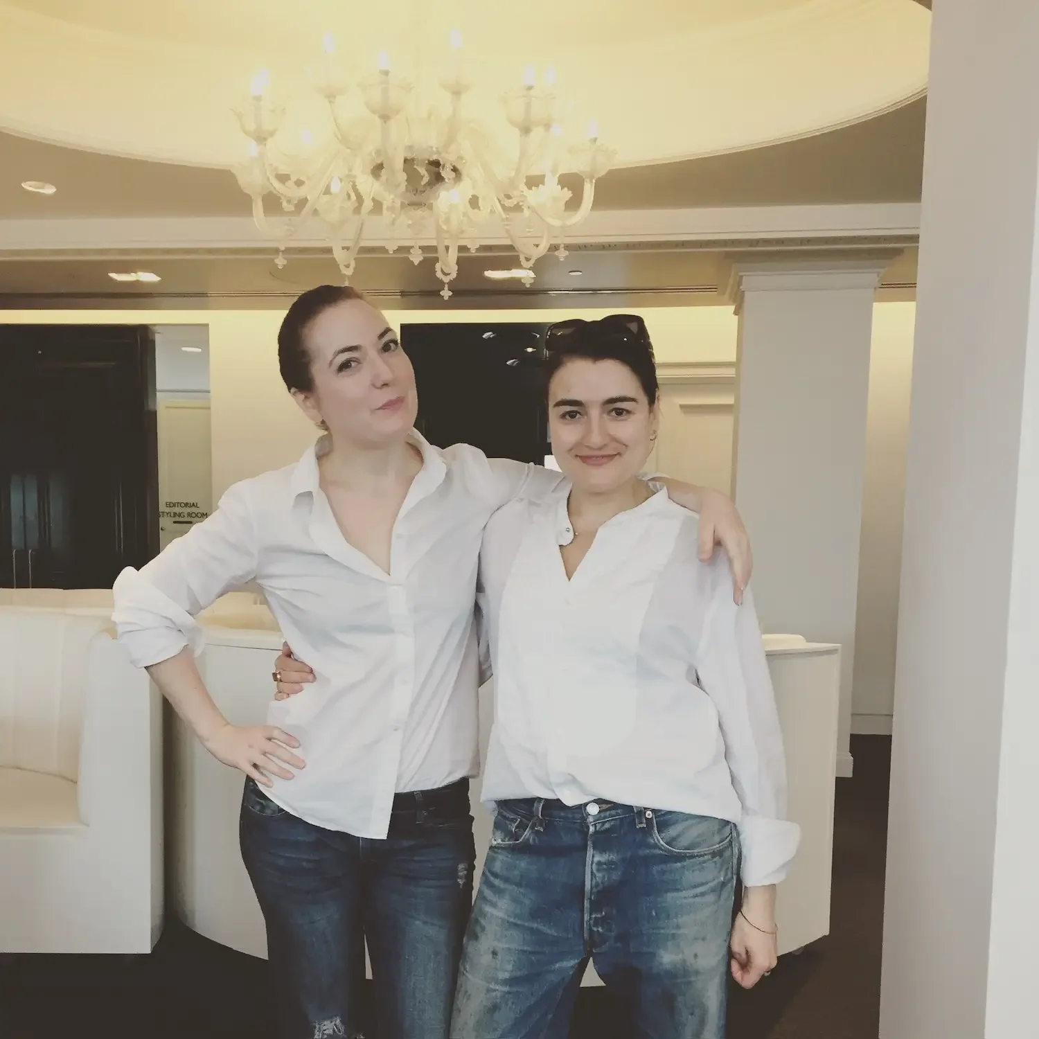 Two women standing side by side in a hotel lobby, wearing white shirts and jeans, smiling with arms around each other.