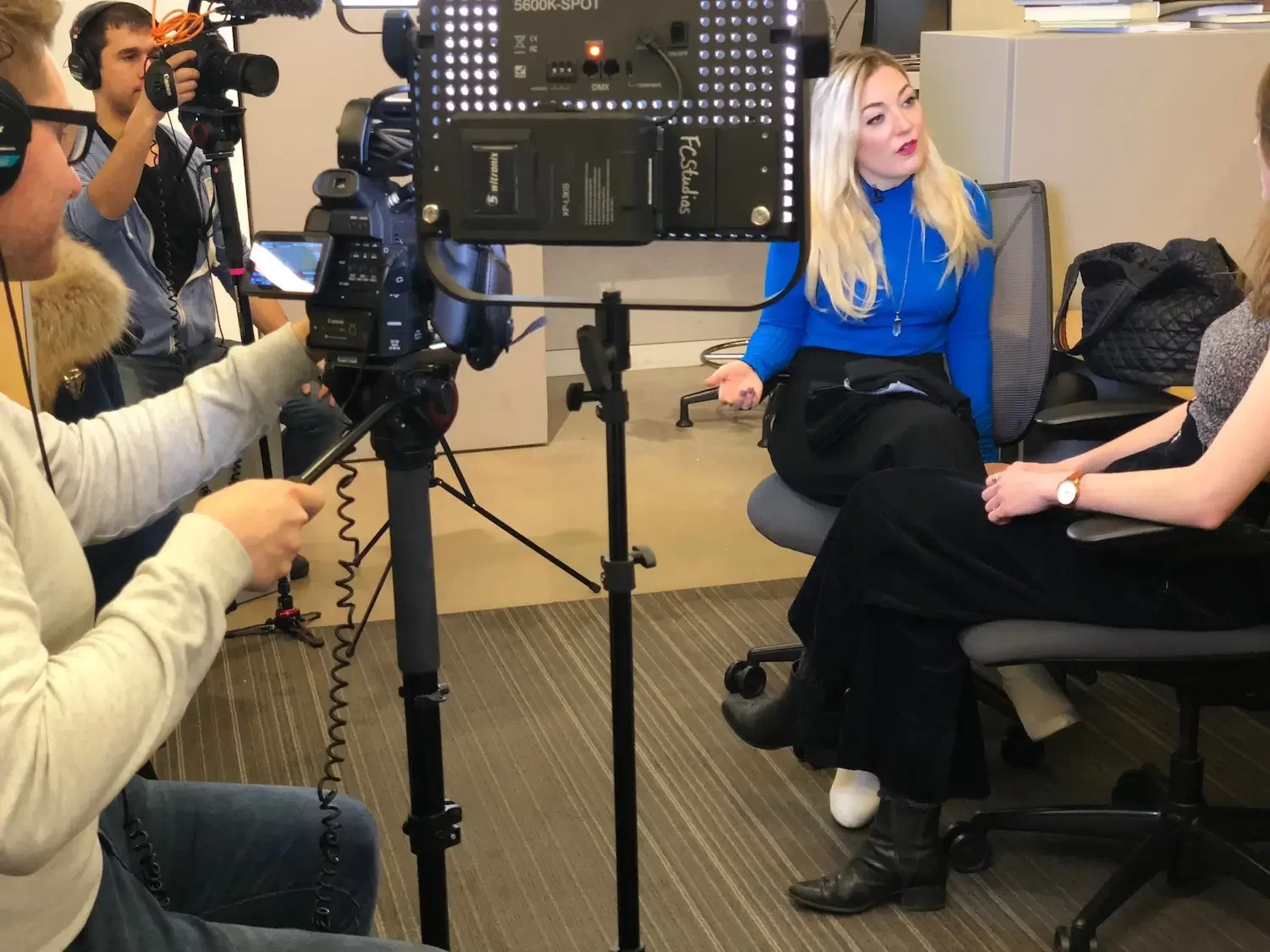 A woman in a blue sweater being interviewed by another woman while a camera crew films the scene in an office setting.