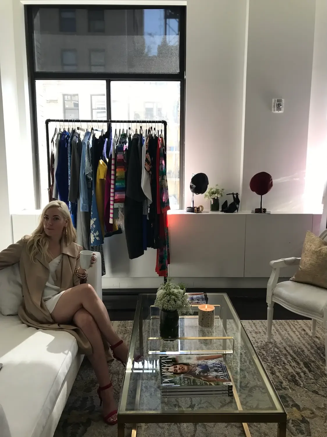 A woman is sitting on a white sofa holding a mug in a living room with a glass coffee table, a clothing rack with colorful clothes, and a window in the background.