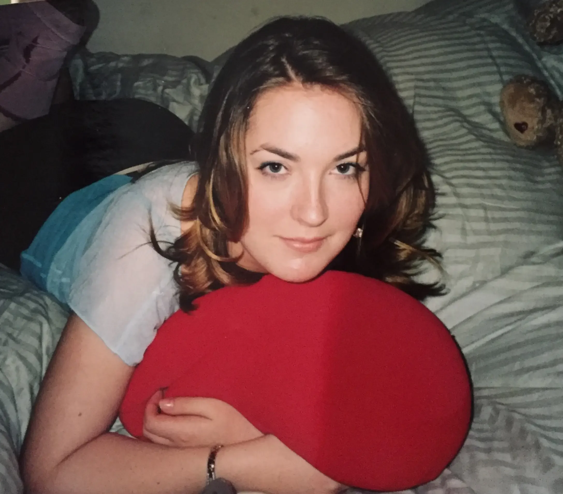 Young woman with brown hair and fair skin lying on a bed, hugging a red pillow, looking at the camera with a slight smile.