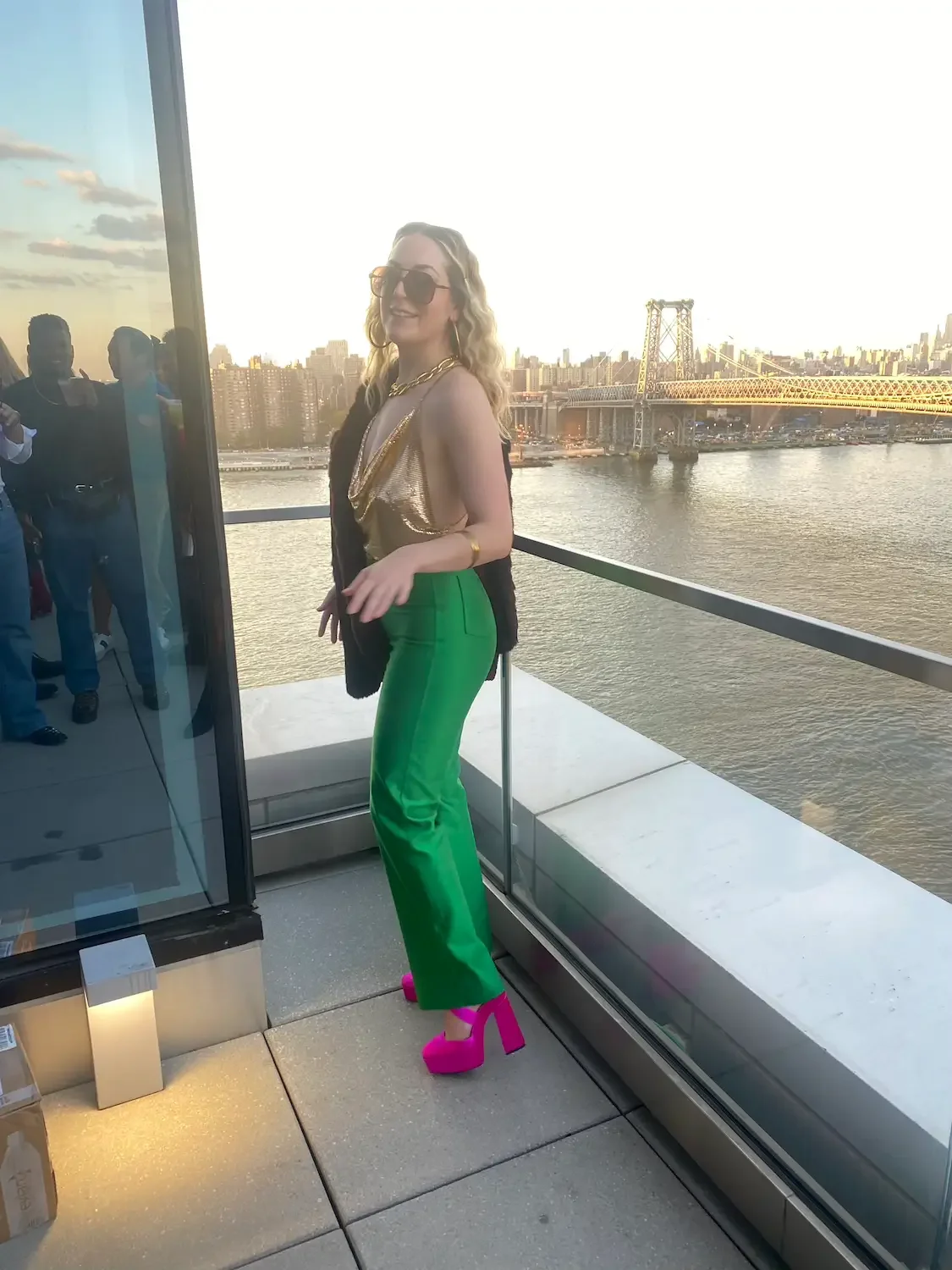 Nicole Russo wearing large sunglasses, gold top, black blazer, green pants, and bright pink platform shoes standing on a rooftop balcony with a city skyline and bridge in the background during sunset.